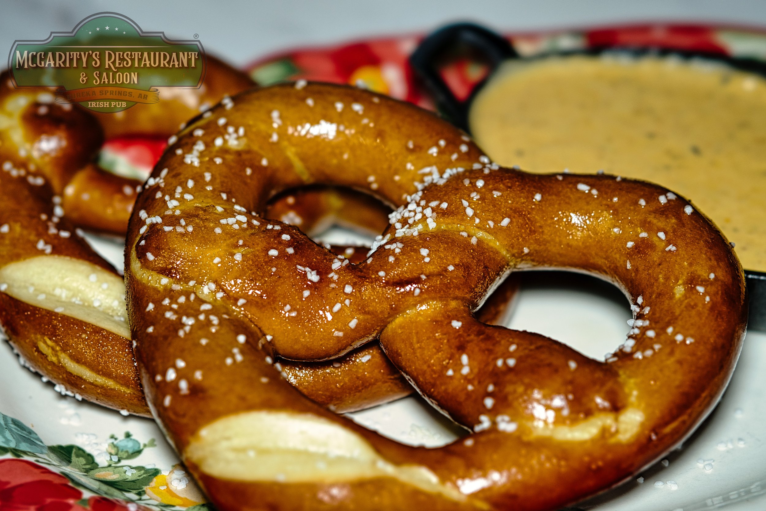 Close-up of a salted soft pretzel with coarse salt, served on a white plate with a side of mustard dip and a condiment container in the background.
