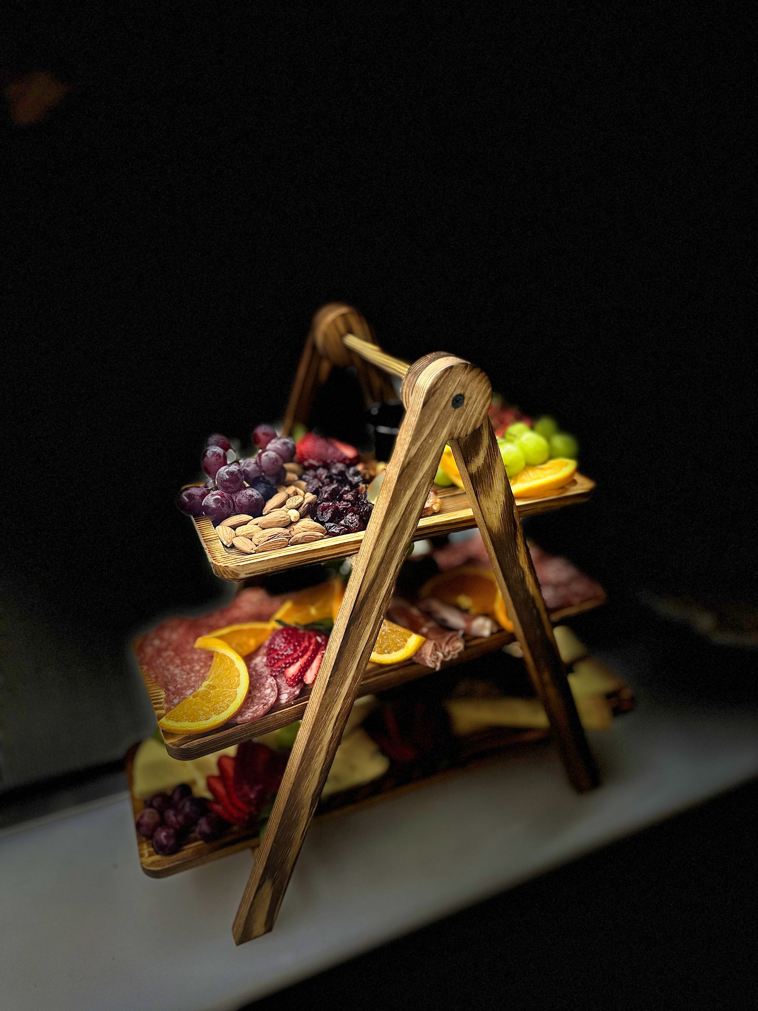Wooden three-tiered serving tray with assorted fruits, nuts, and meats on a white surface against a dark background.