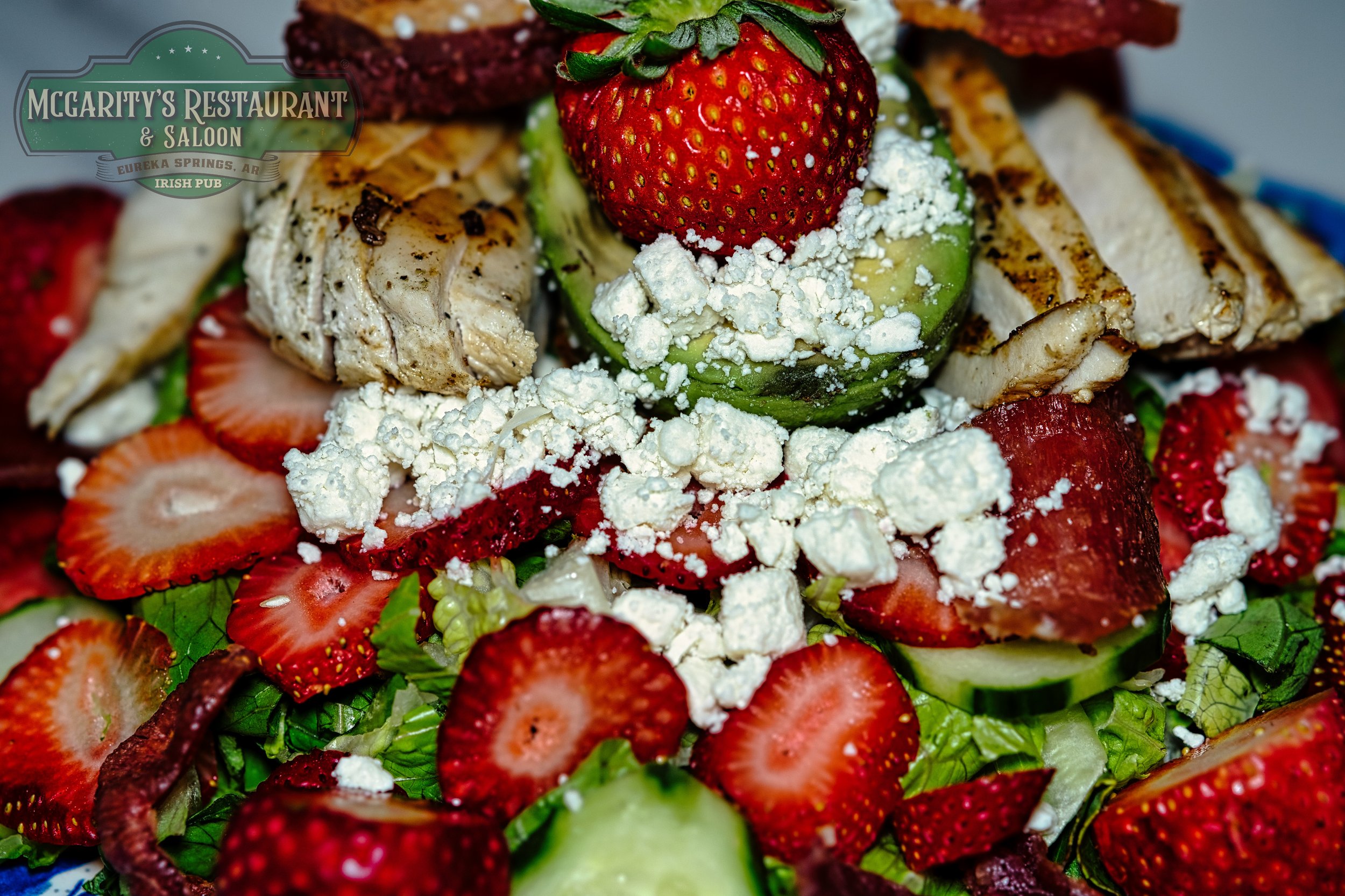 Close-up of a fresh strawberry and cucumber salad topped with grilled chicken pieces, crumbled feta cheese, and sliced strawberries, with a McGarity's Restaurant & Saloon logo in the top left corner.