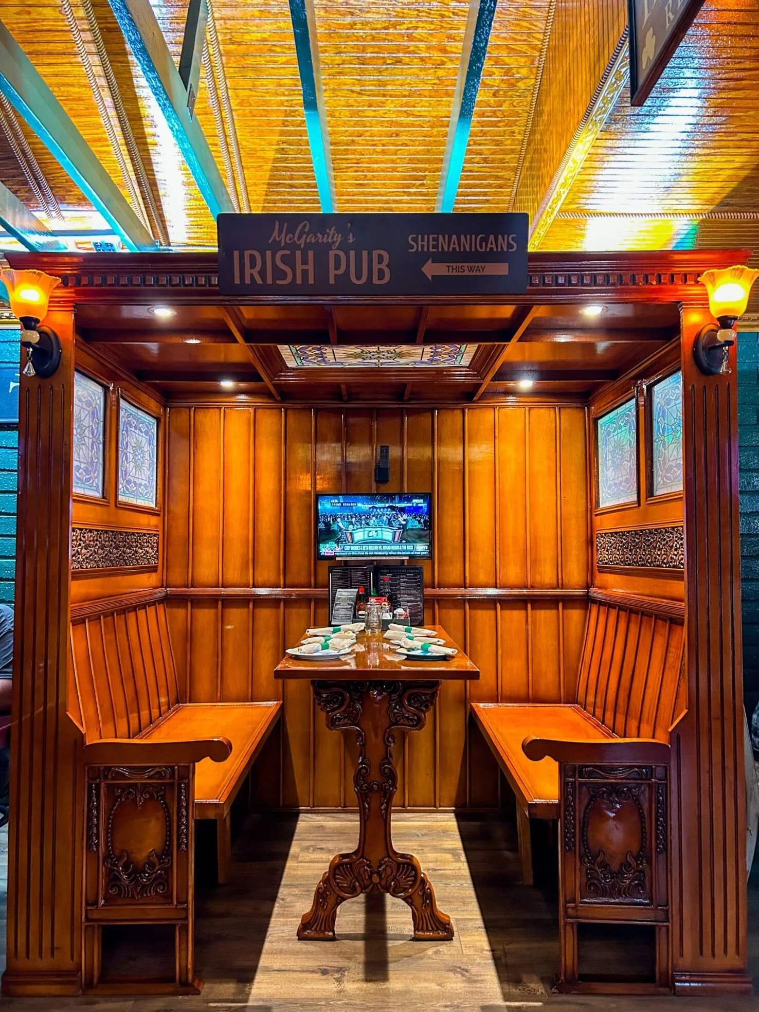 A cozy Irish pub booth with wooden paneling, a table set for four with napkins and glasses, a TV screen, and a sign pointing to Shenanigans bar.