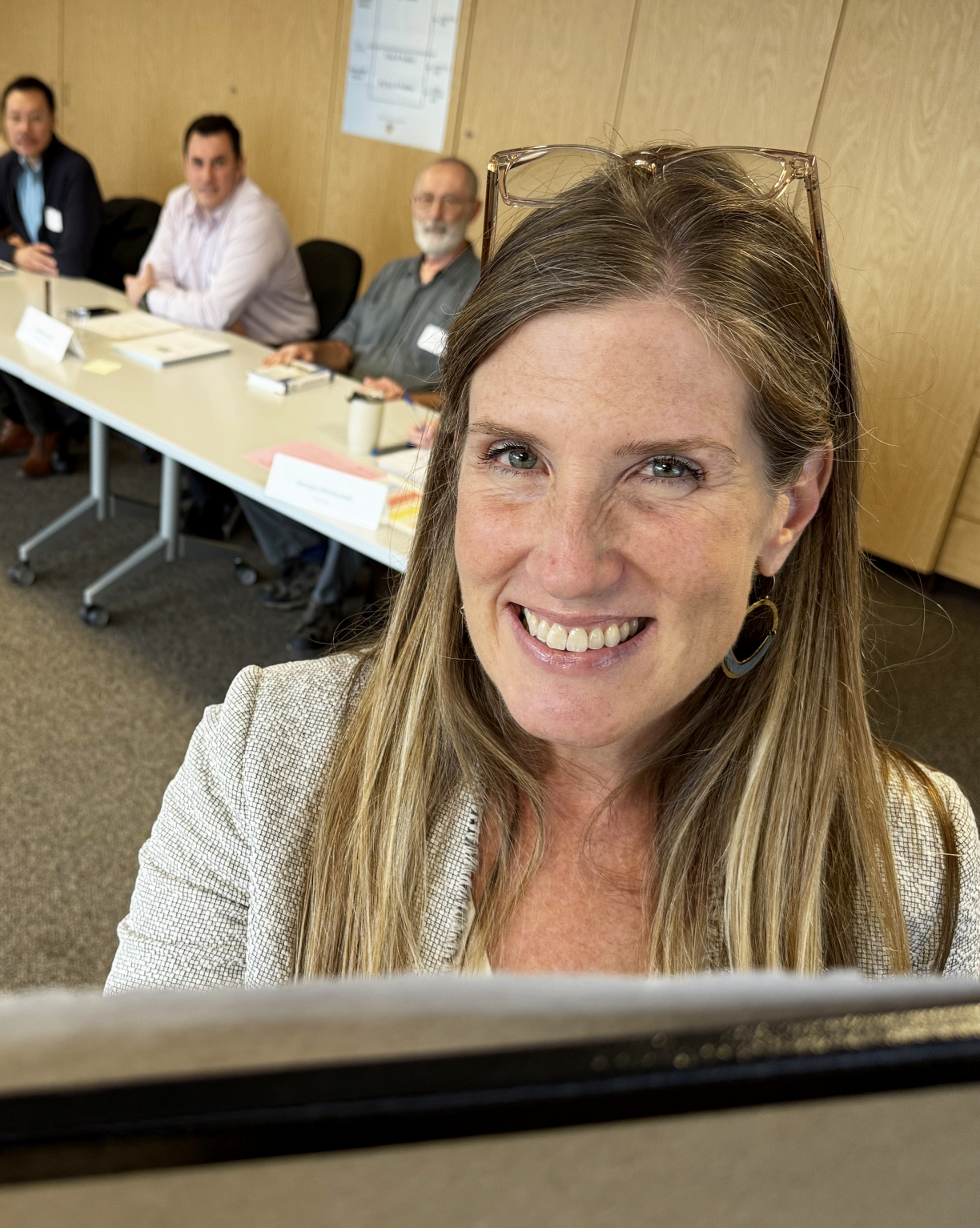 A woman with long light brown hair smiling at the camera, with a conference room and three men sitting at a table in the background.