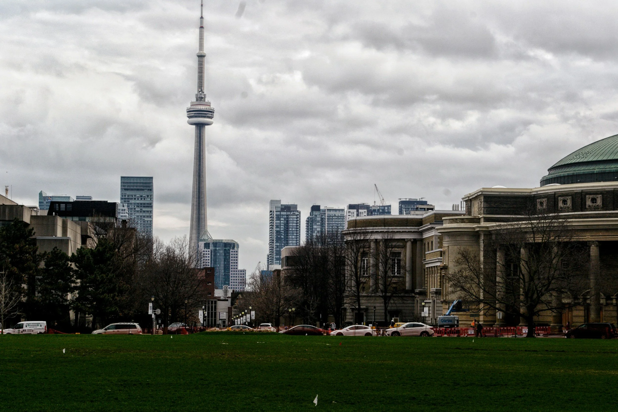 Cityscape view featuring the CN Tower with surrounding high-rise buildings on a cloudy day, seen from a grassy park with trees and parked cars in the foreground.
