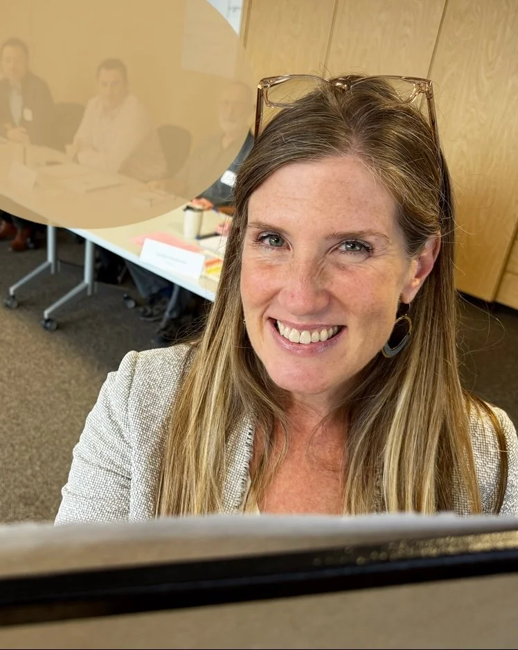 A woman with long blond hair, glasses on her head, smiling at the camera, in a professional setting with a conference room in the background.