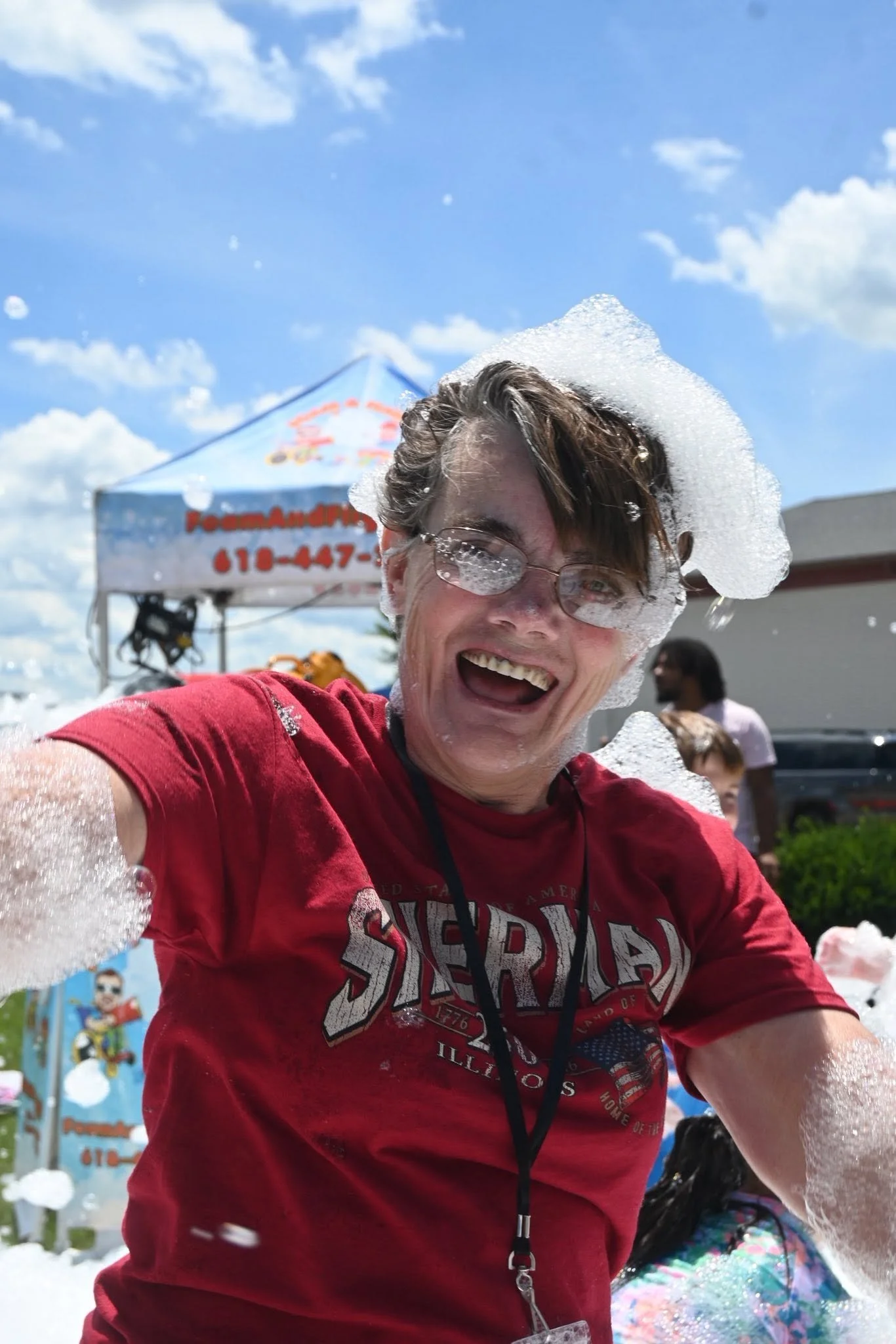 A person smiling and covered in soap foam in an outdoor setting, with a blue sky and clouds overhead. They are wearing glasses and a red t-shirt, with a booth in the background.