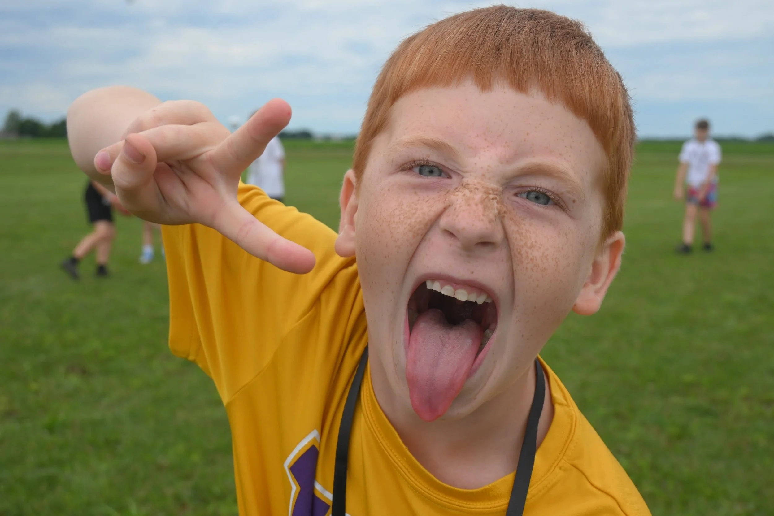A young boy with red hair and freckles, making a silly face with his mouth open and tongue out, pointing towards the camera, on a grassy field with blurred children in the background.