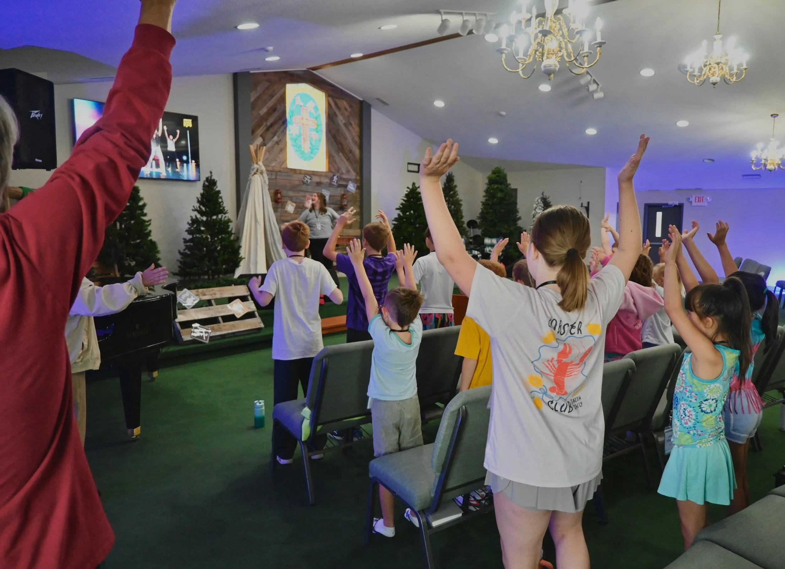 Children raising their hands during a church service in a decorated room with Christmas trees, a cross on a wooden wall, and chandeliers.