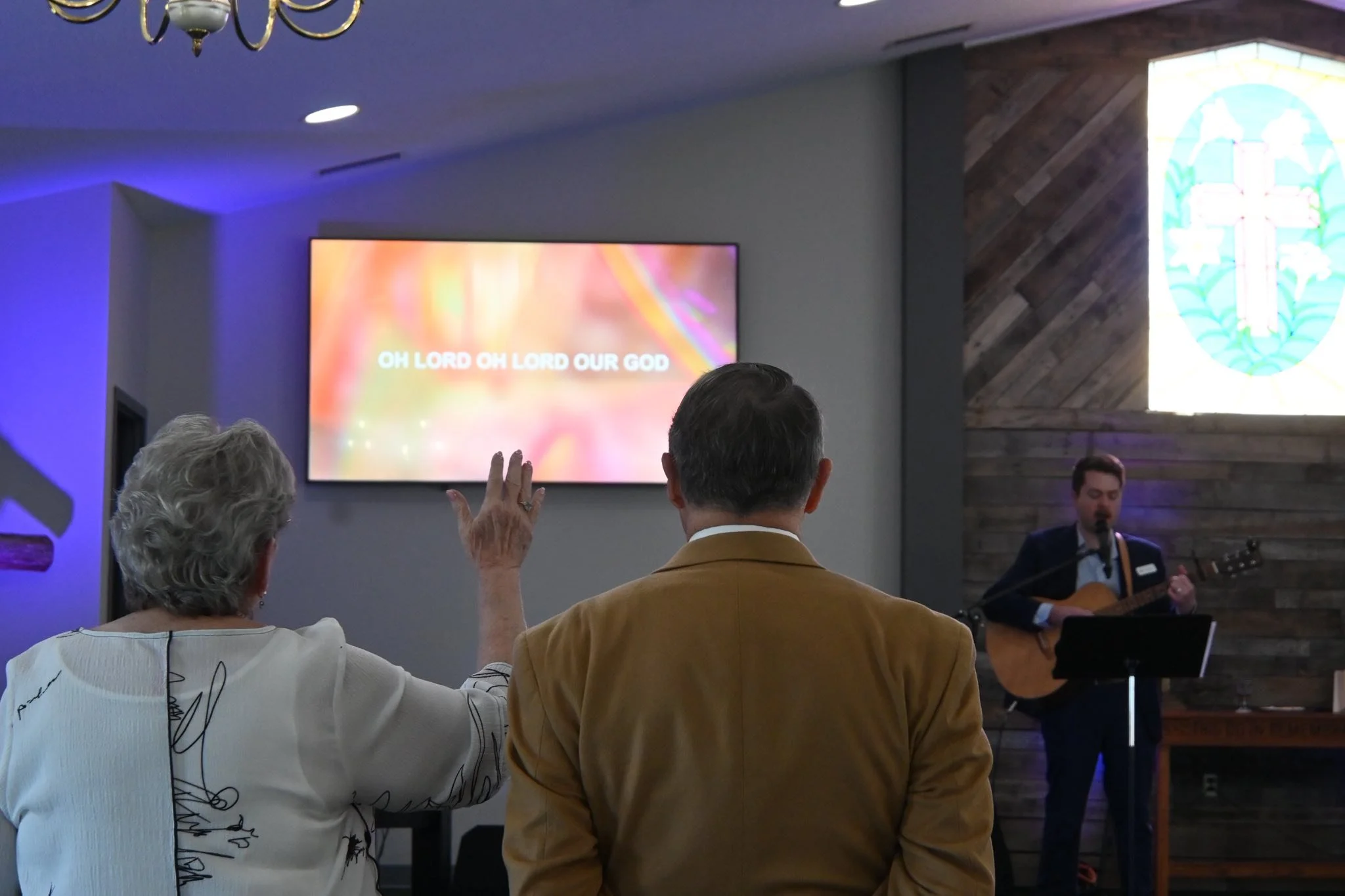 A church service or gathering with people seated, singing or praying, with a man playing guitar and a preacher or pastor reading from a book. The screen behind displays the words 'OH LORD OH LORD OUR GOD'. There is a stained glass window on the side wall.