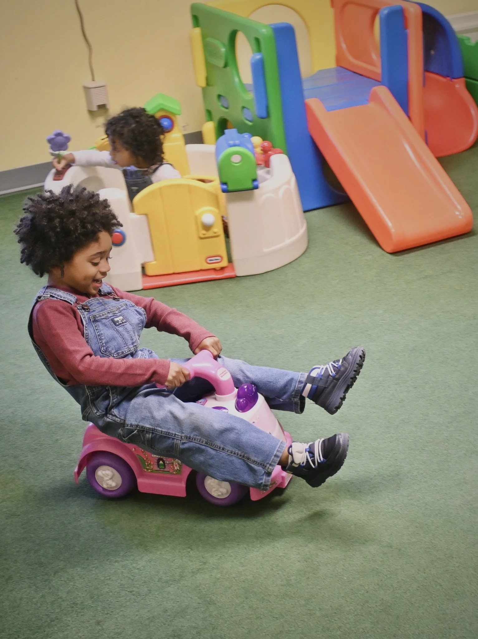 Two young children playing and smiling inside a playroom with a green carpet. One child is riding a pink battery-powered toy car, and the other is playing in a colorful plastic playhouse with a slide in the background.