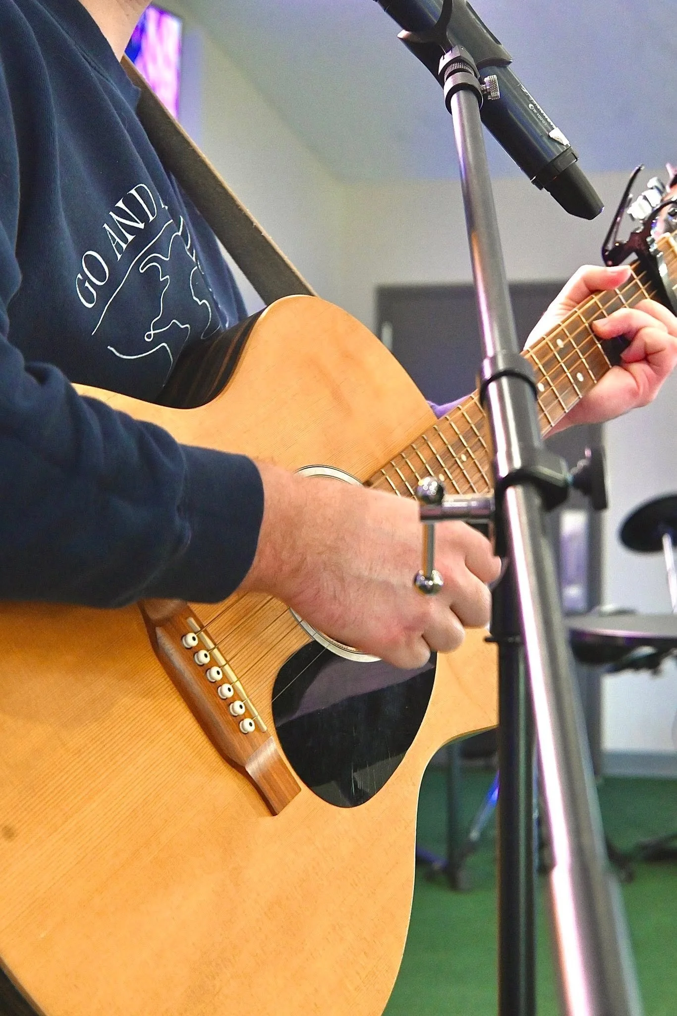 Close-up of a person playing an acoustic guitar, with their left hand on the fretboard and a microphone stand in the foreground.
