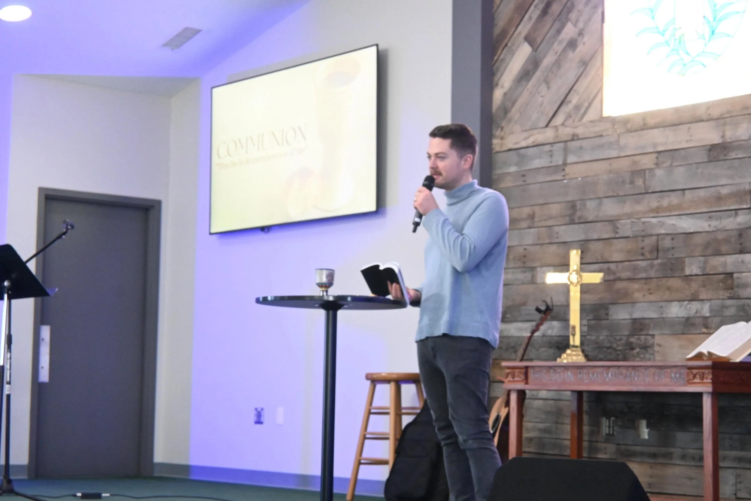 A man in a light blue sweater holding a microphone and a book, standing behind a round table with a glass and a small object on it, inside a church with wooden wall panels, a cross, and a Bible stand in the background.