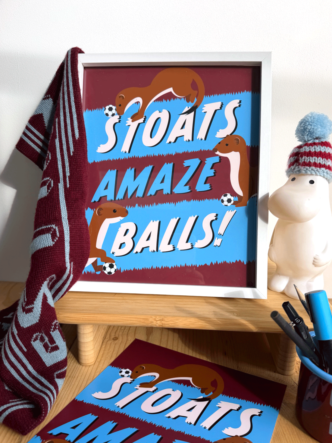 Framed poster in West Ham claret and blue colourswith playful graphic design featuring stoats and footballs, reading 'Stoats Amaze Balls!', placed on a wooden desk with a moomin wearing a mini football hat and office supplies around it.