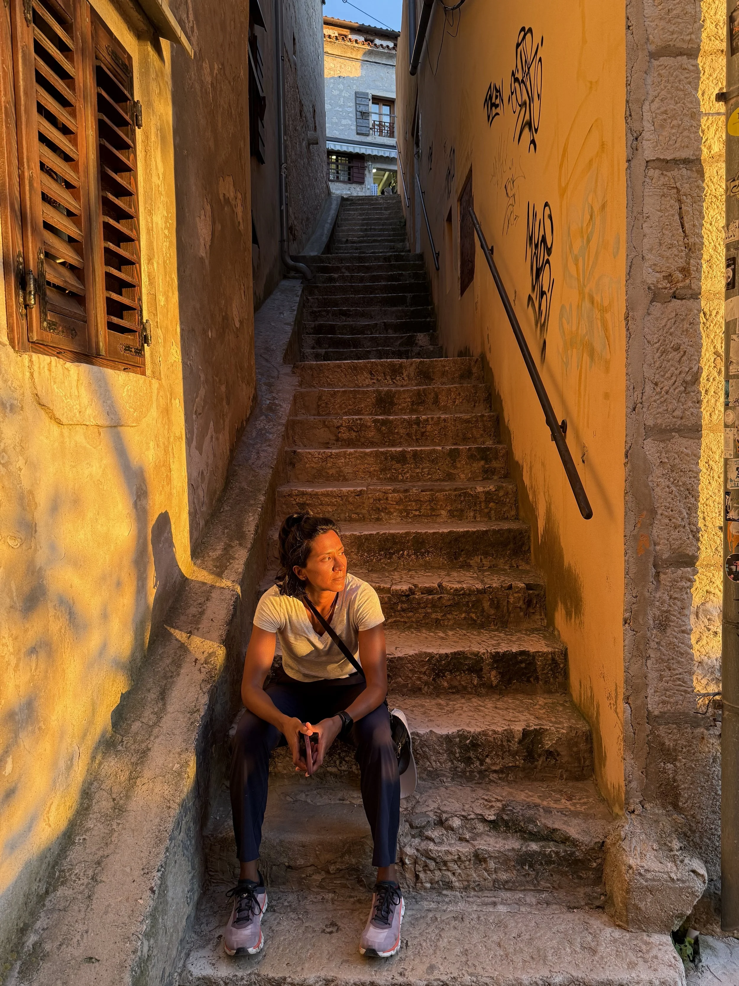 A woman sitting on a step in a narrow alleyway with stairs leading upward. She is wearing a white shirt, dark pants, and sneakers, holding a phone in her hands. The alley has yellow and gray walls, with graffiti on the right wall and a window on the left wall.