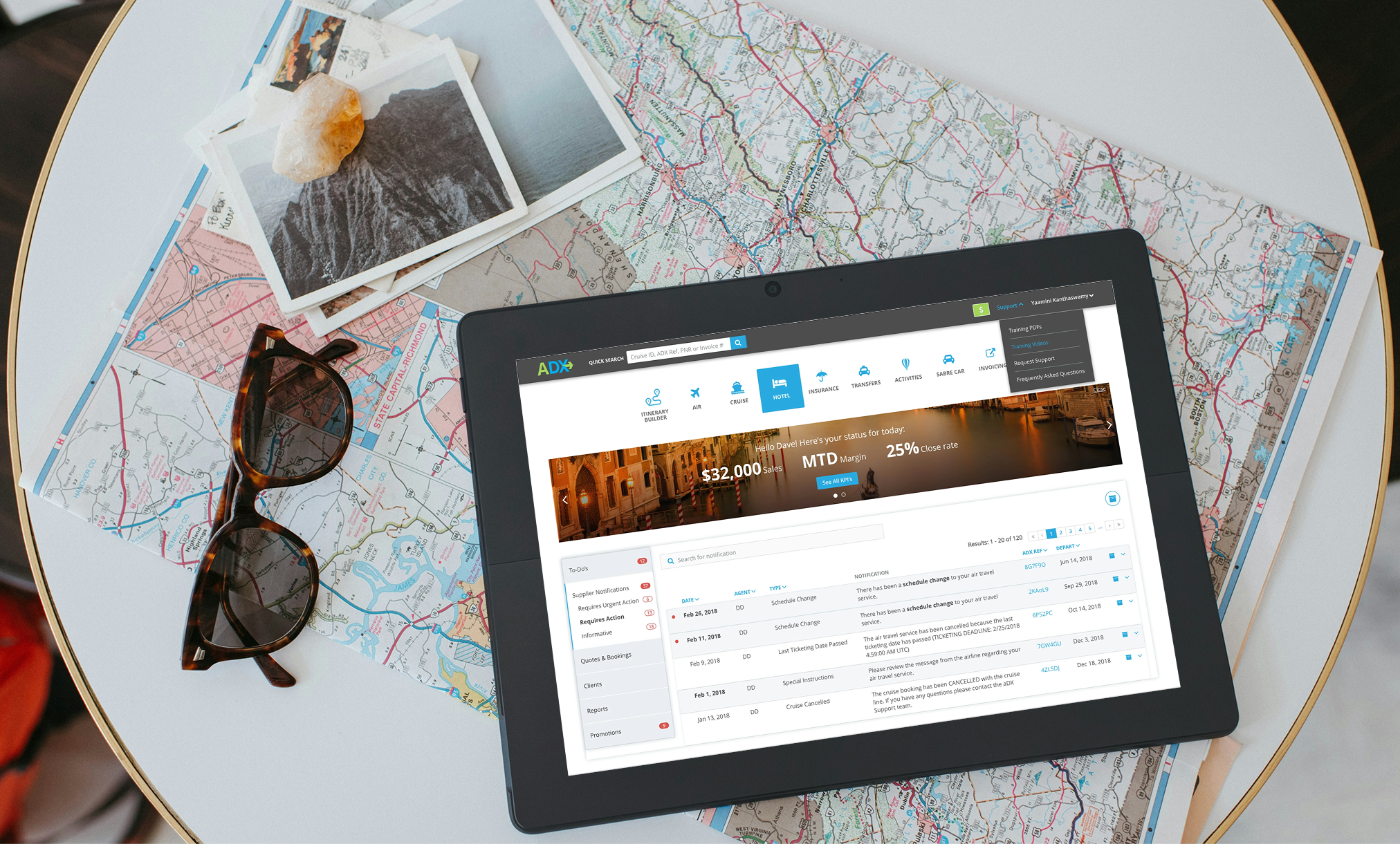 Table with maps, photographs, and a digital device displaying a travel booking website. A pair of sunglasses rests on the maps, which include a detailed road map and photographs of mountains and water.