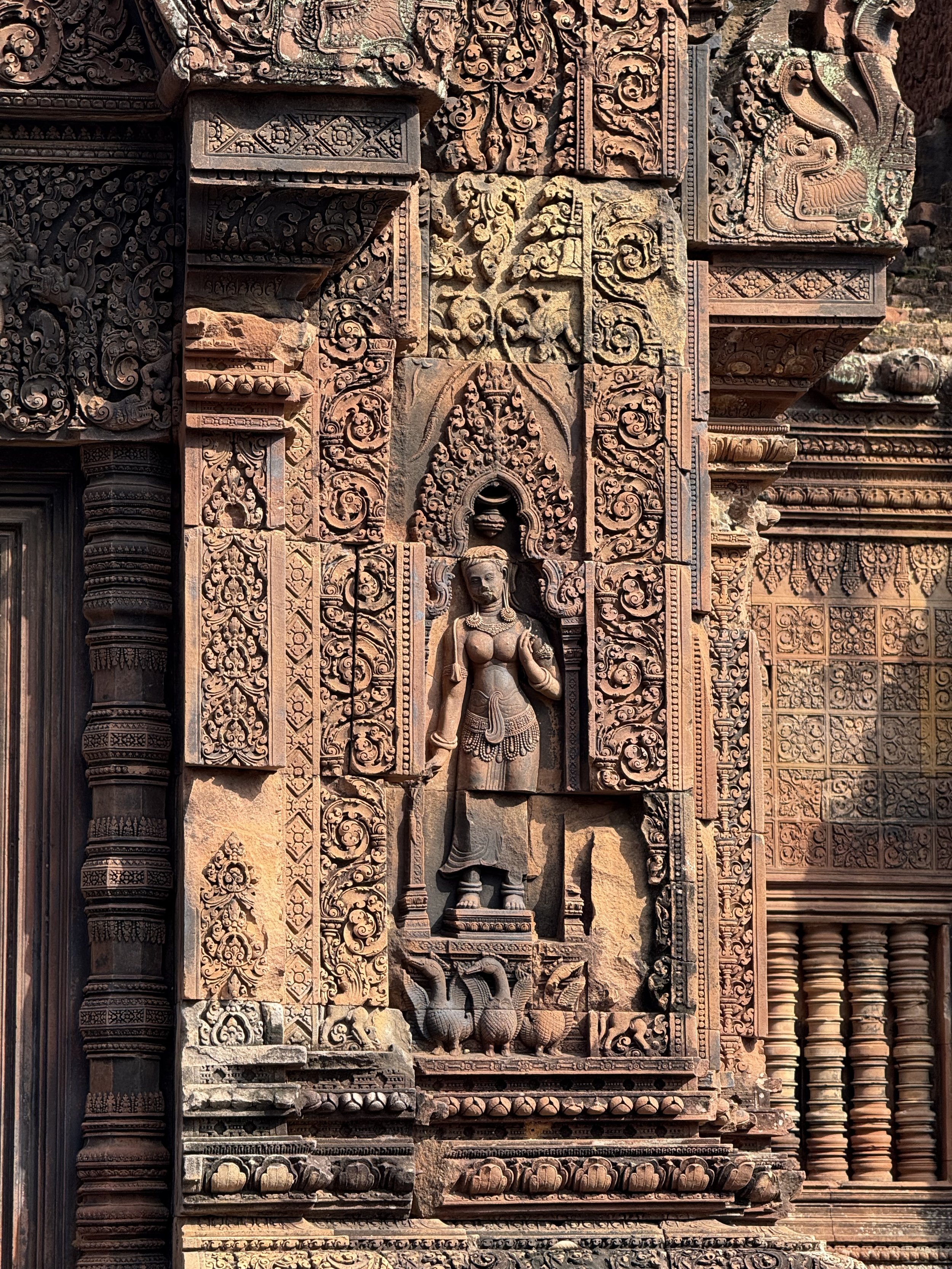 Ancient stone wall carved with intricate patterns and figures, including a standing female figure with elaborate jewelry, surrounded by decorative motifs and architectural details.