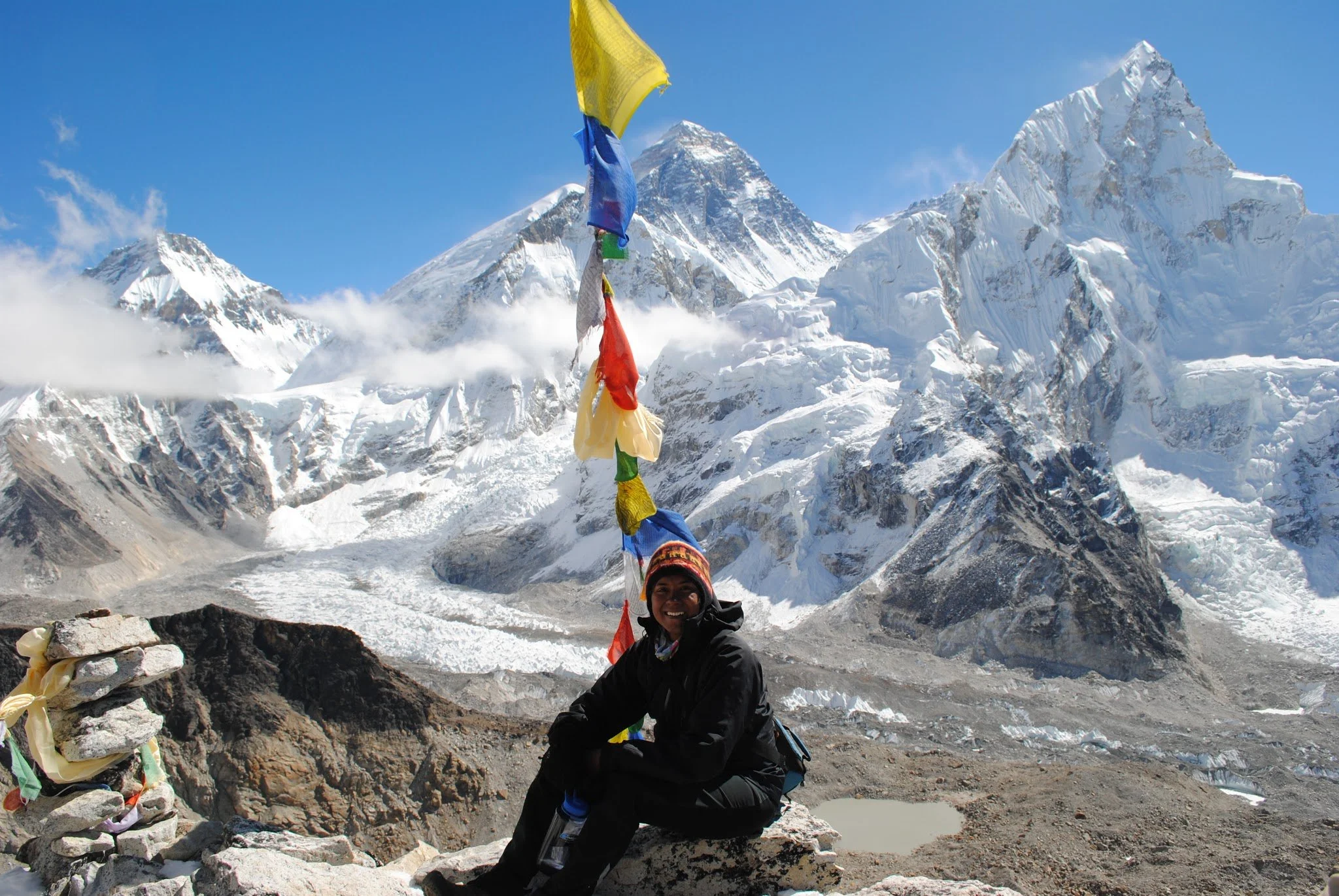 A person smiling and wearing a black jacket and a colorful hat, sitting on rocks in a snowy mountain landscape with high peaks and prayer flags.