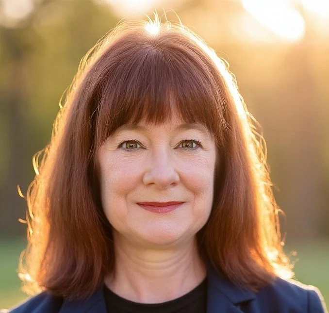 A middle-aged woman with shoulder-length reddish-brown hair smiling outdoors during sunset.