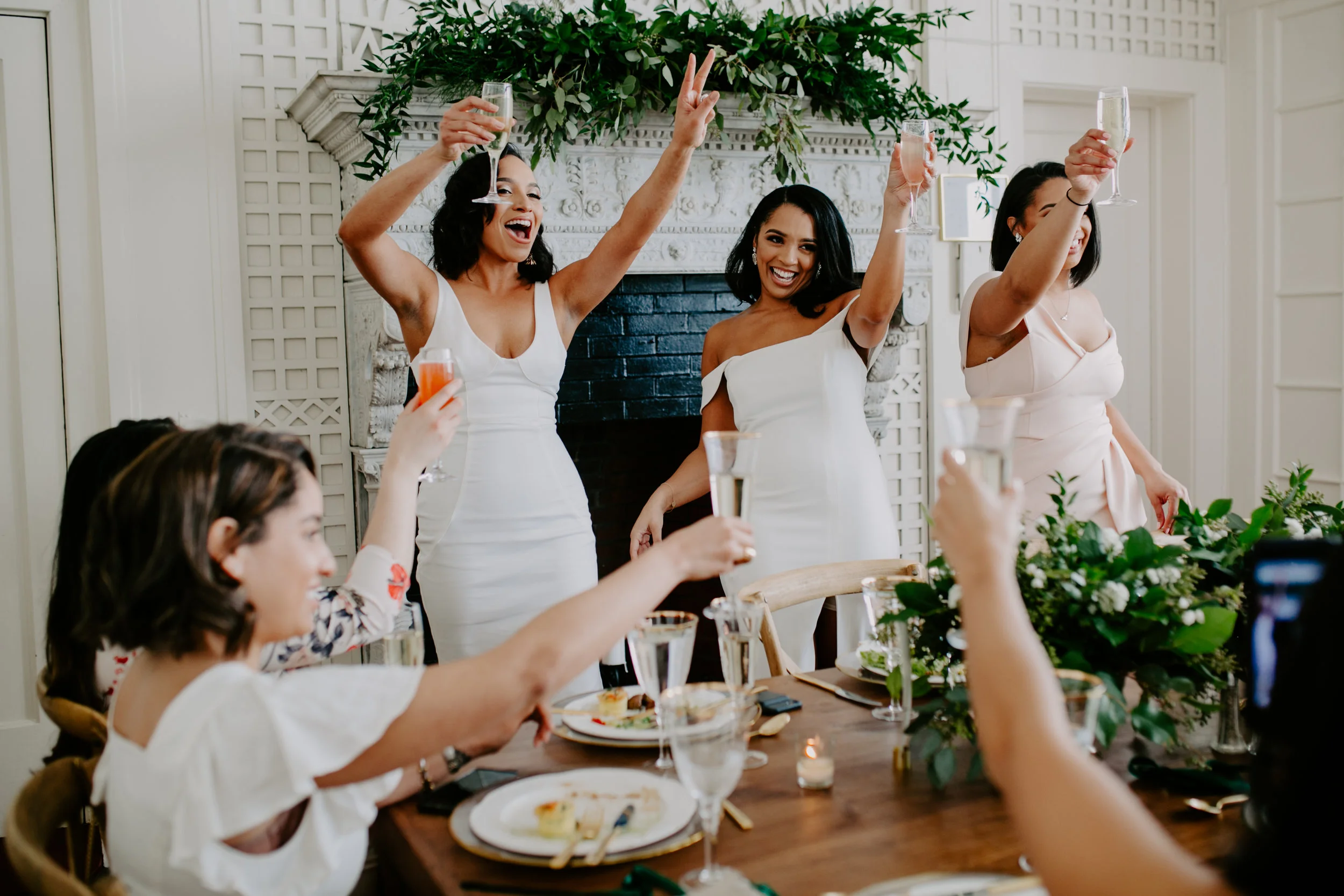 Women celebrating at a wedding reception, raising glasses for a toast, seated and standing around a decorated table with food and drinks.