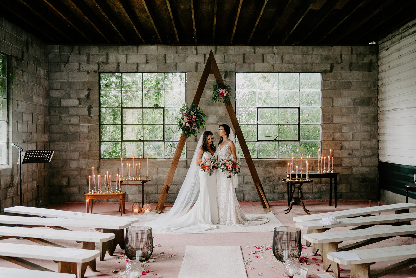 Two brides in wedding gowns standing inside an industrial-style venue with large windows, floral arrangements, and lit candles, under a wooden triangular arch decorated with flowers and greenery.