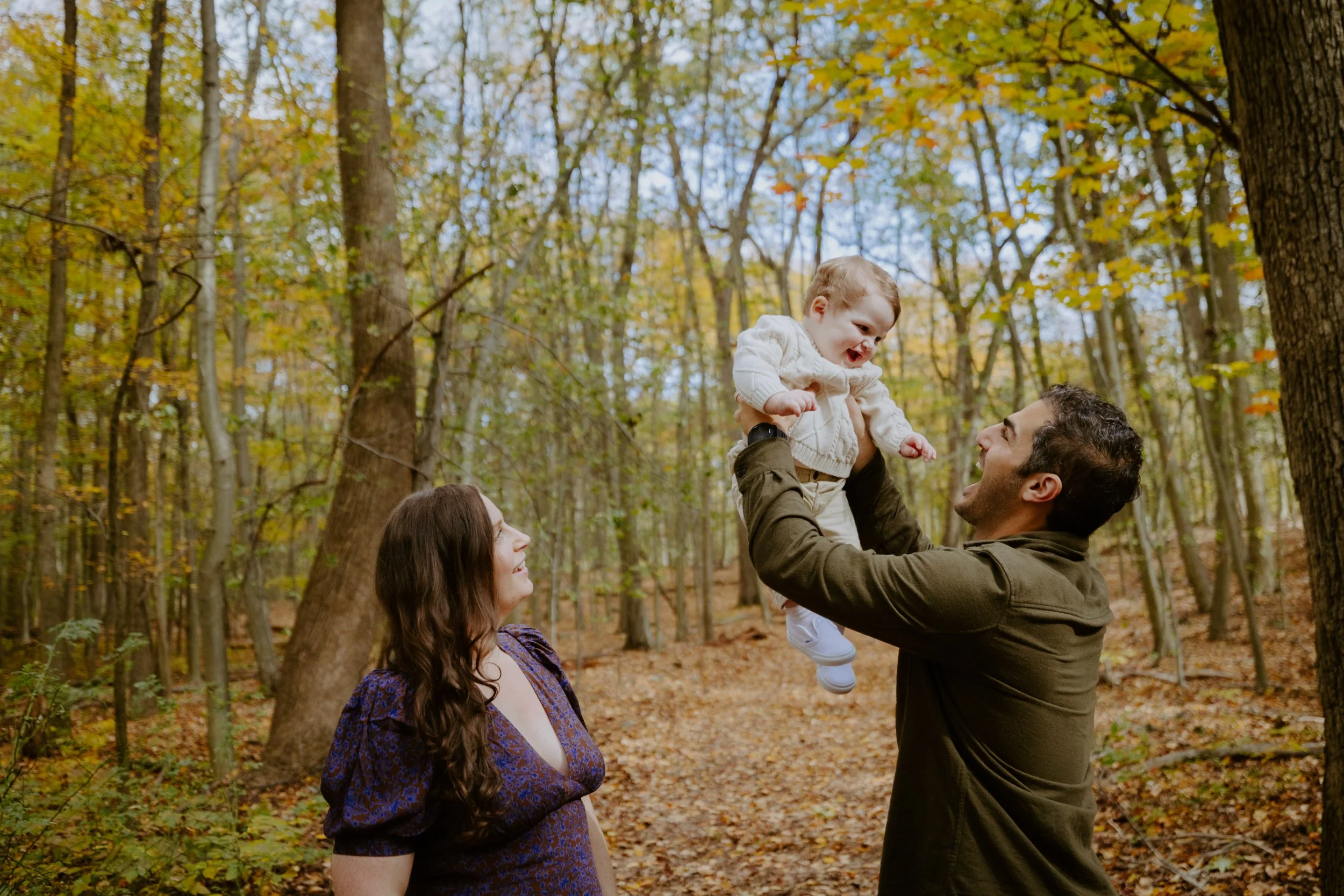 A family of three outdoors in a forest during fall, with the father lifting a smiling child into the air while the mother watches nearby.