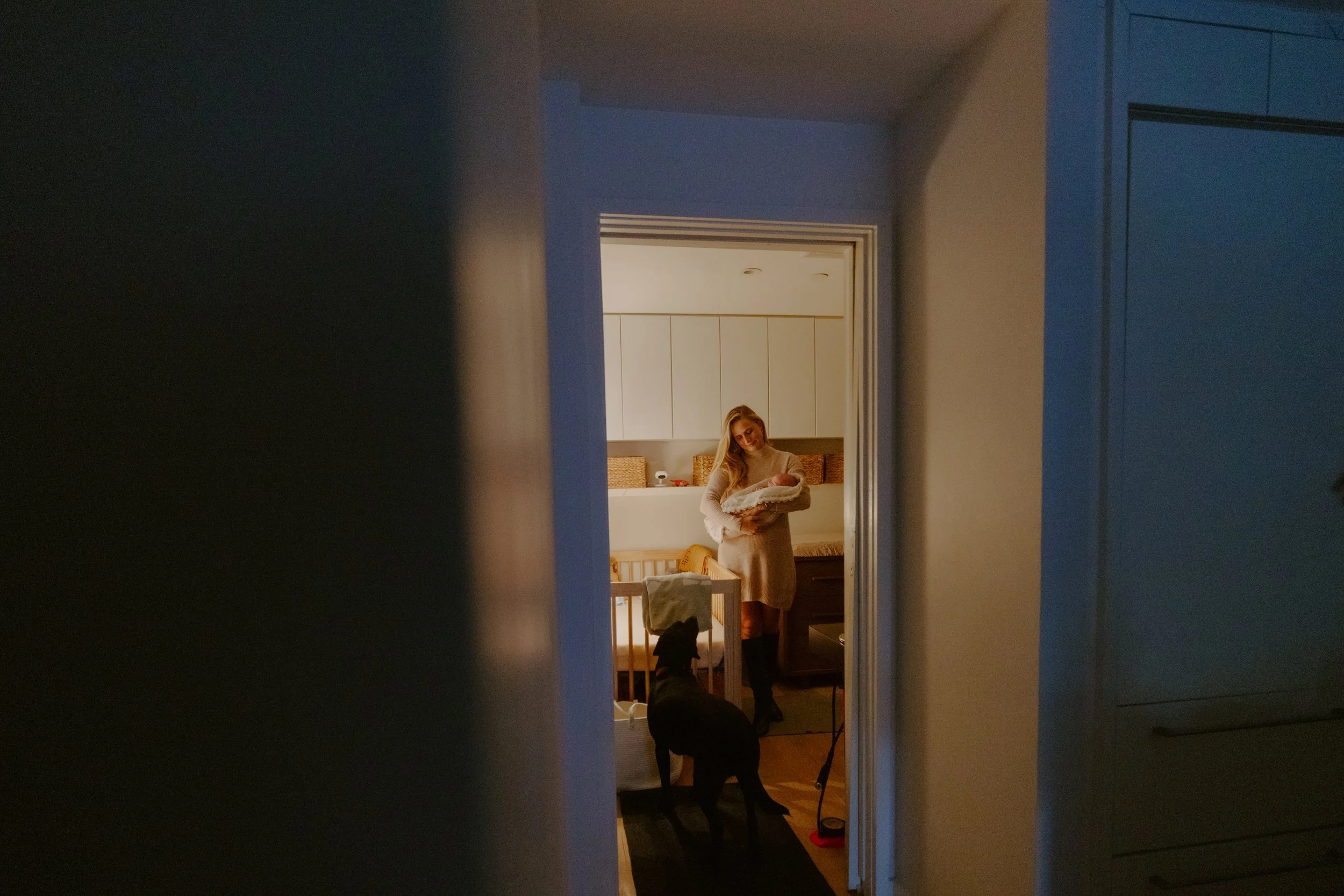 A woman holding a baby in a baby crib seen through a doorway, with a black dog standing on a mat in the room.
