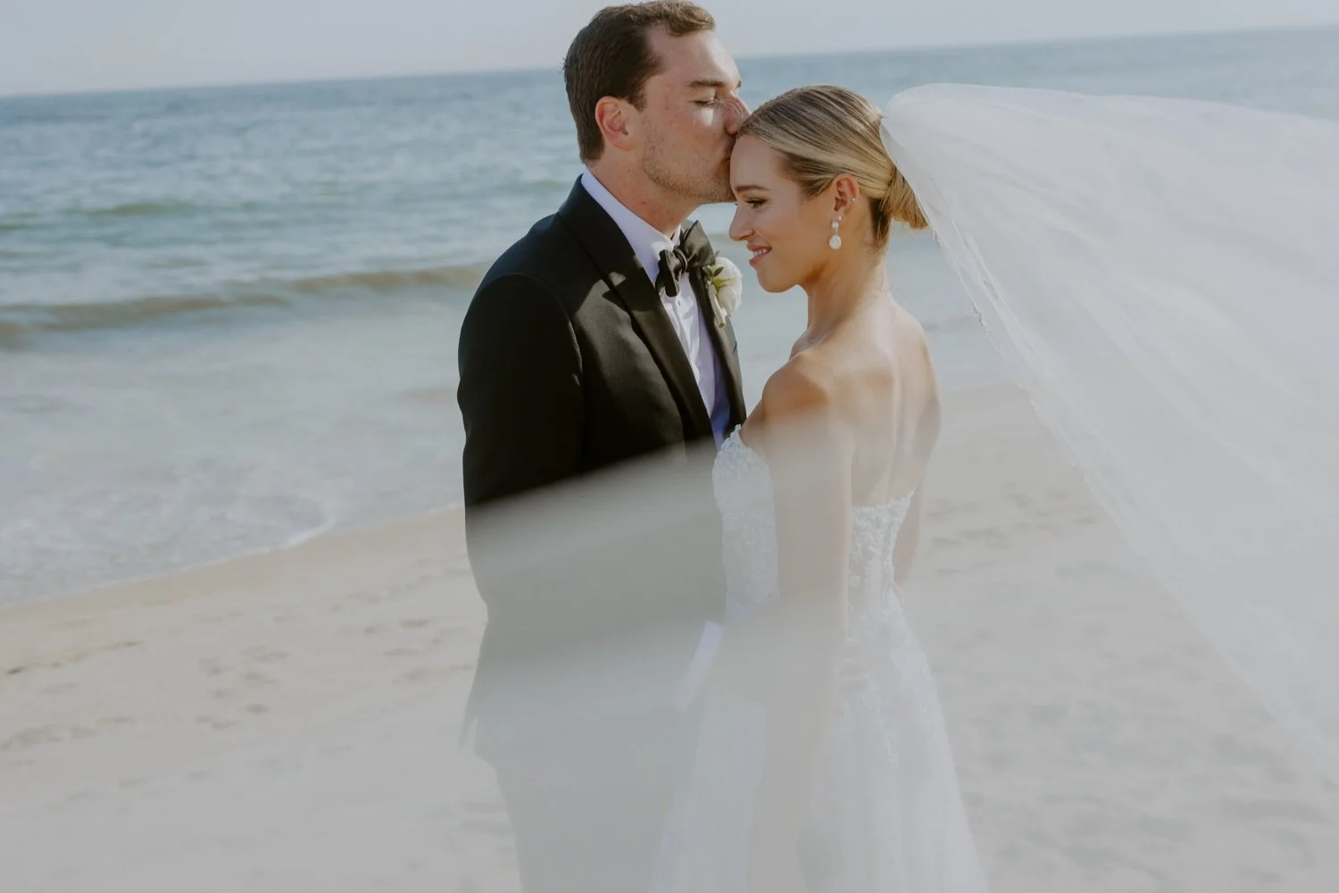 A bride and groom standing on the beach during their wedding, with the groom kissing the bride's forehead and the ocean in the background.