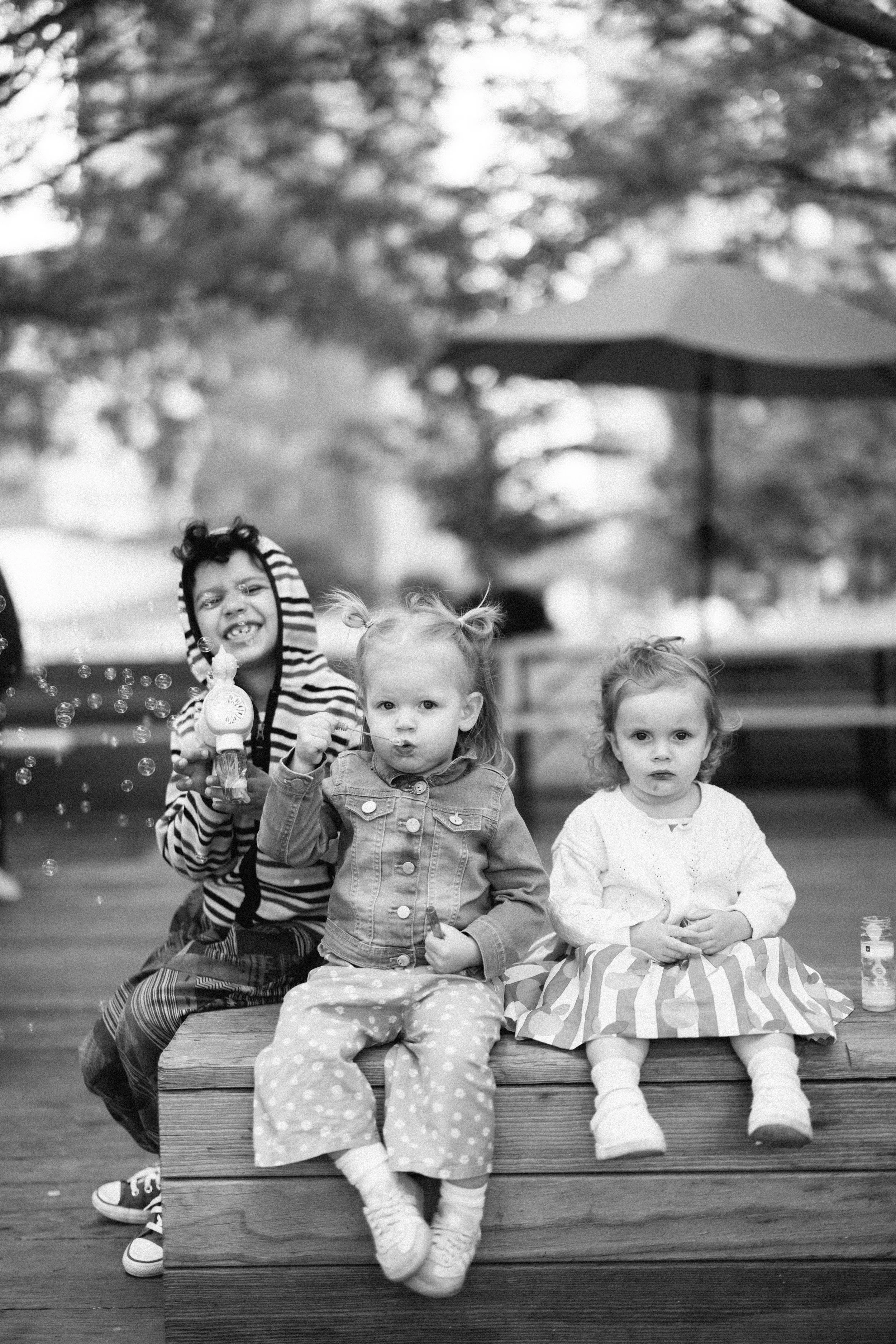 Three young children sitting on a wooden bench outdoors; one girl is blowing bubbles, the other two girls are sitting with neutral expressions, surrounded by trees and a large patio umbrella in the background.