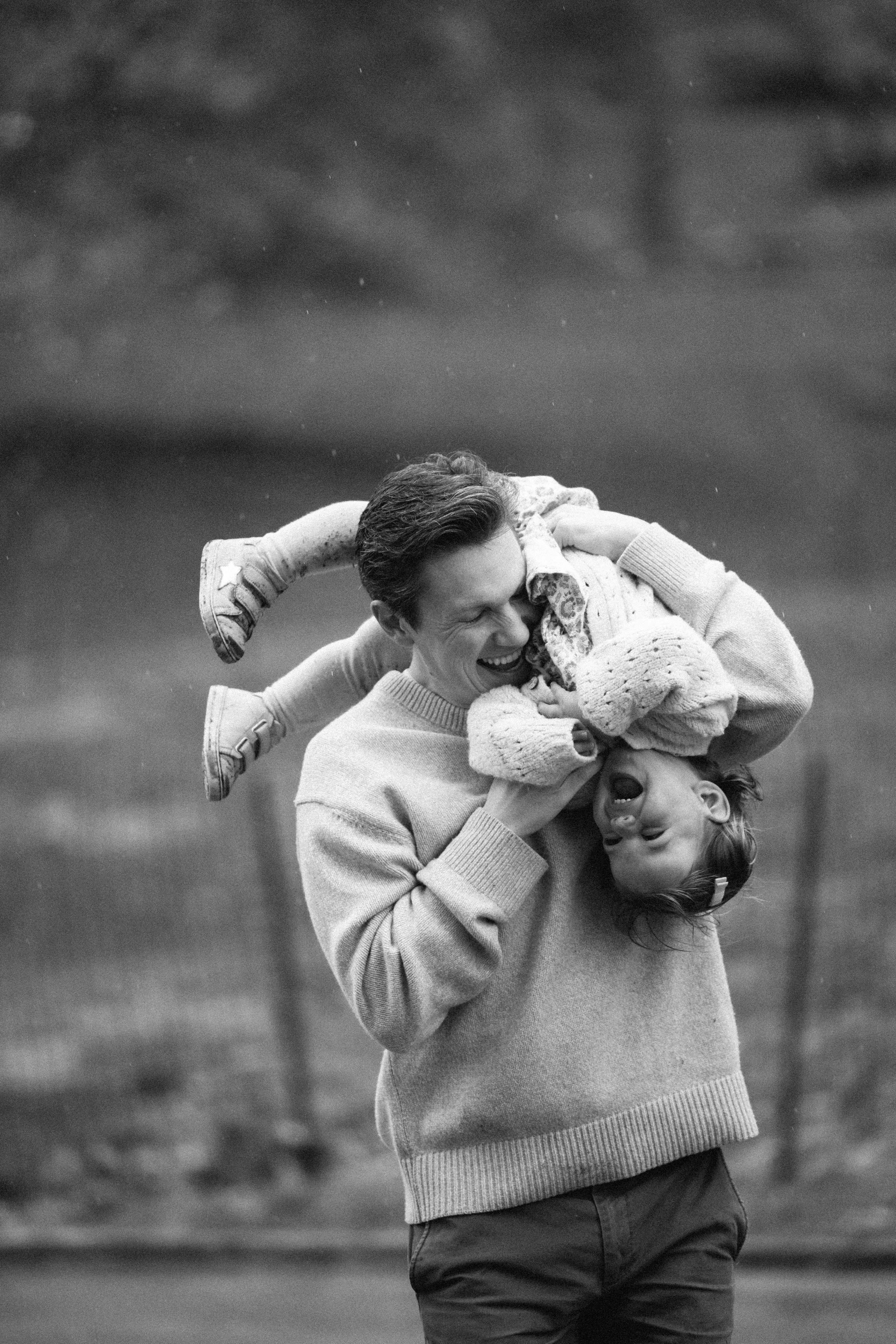 A man with dark hair and a sweater plays with his daughter in a park, holding her upside down. The girl has her eyes closed and mouth open, laughing. The background is blurry, showing trees and an overcast sky.