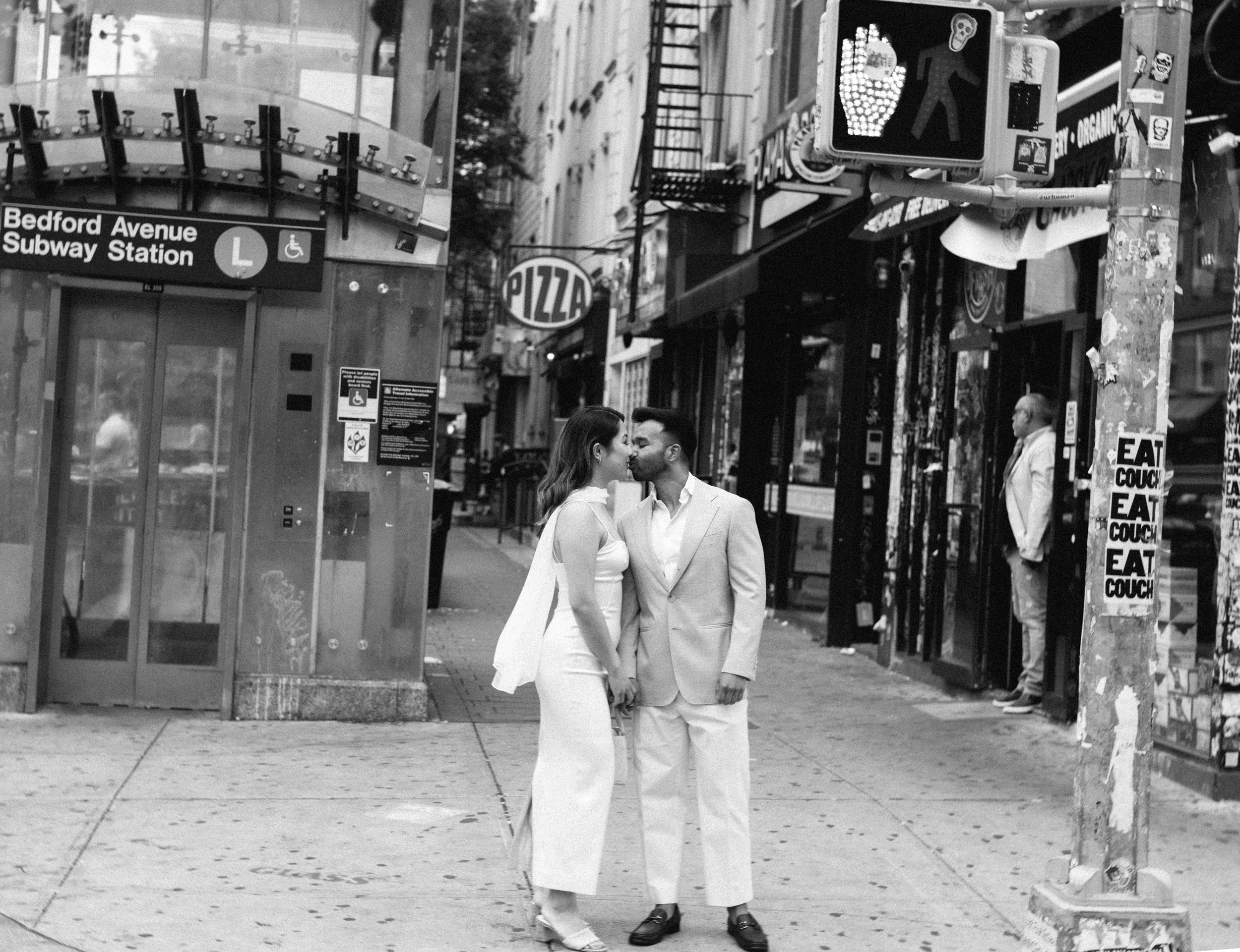A couple dressed in white, holding hands, sharing a kiss on a city sidewalk in front of a subway entrance and a storefront with a pizza sign. There is a pedestrian crossing signal and a man in a light jacket standing nearby.