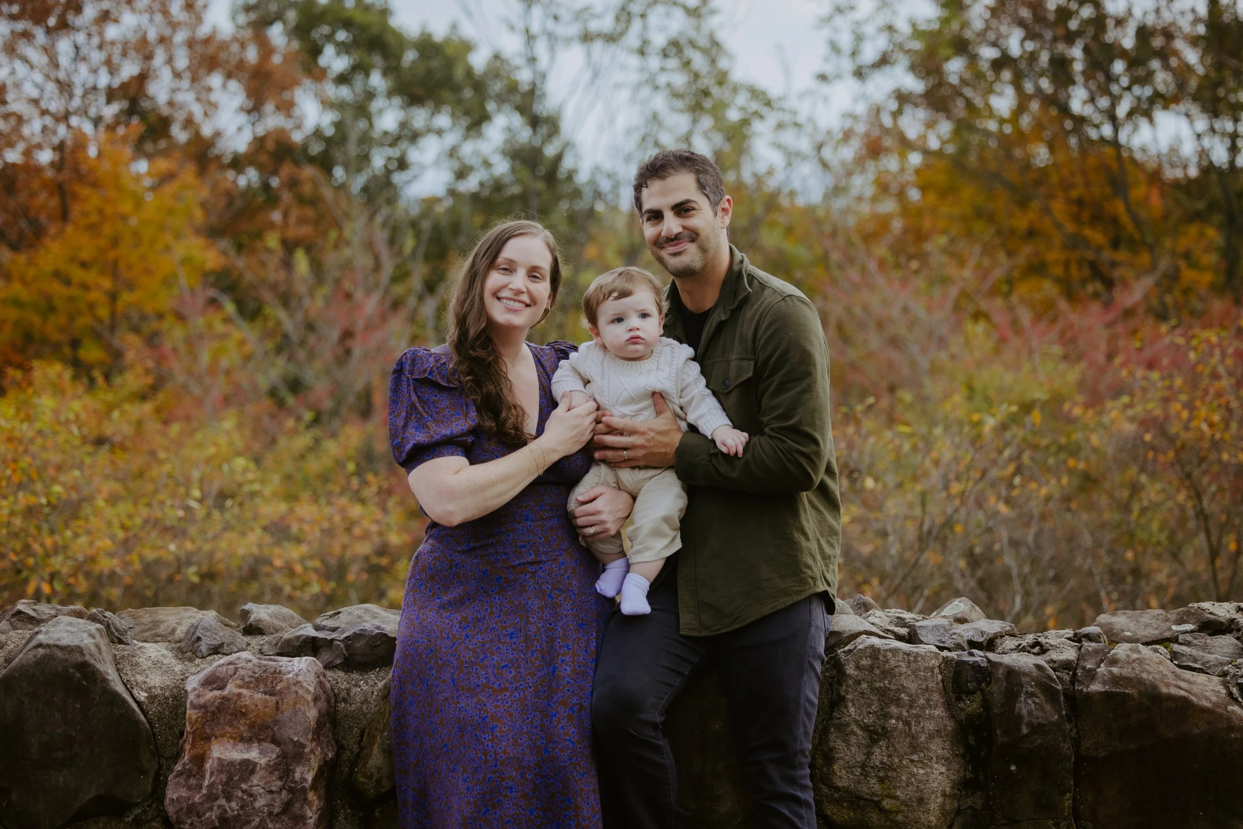 A family of three outdoors in autumn, standing on a stone wall with trees with fall foliage in the background. The woman wears a purple dress, the man in a green jacket, and the child is in light clothing.