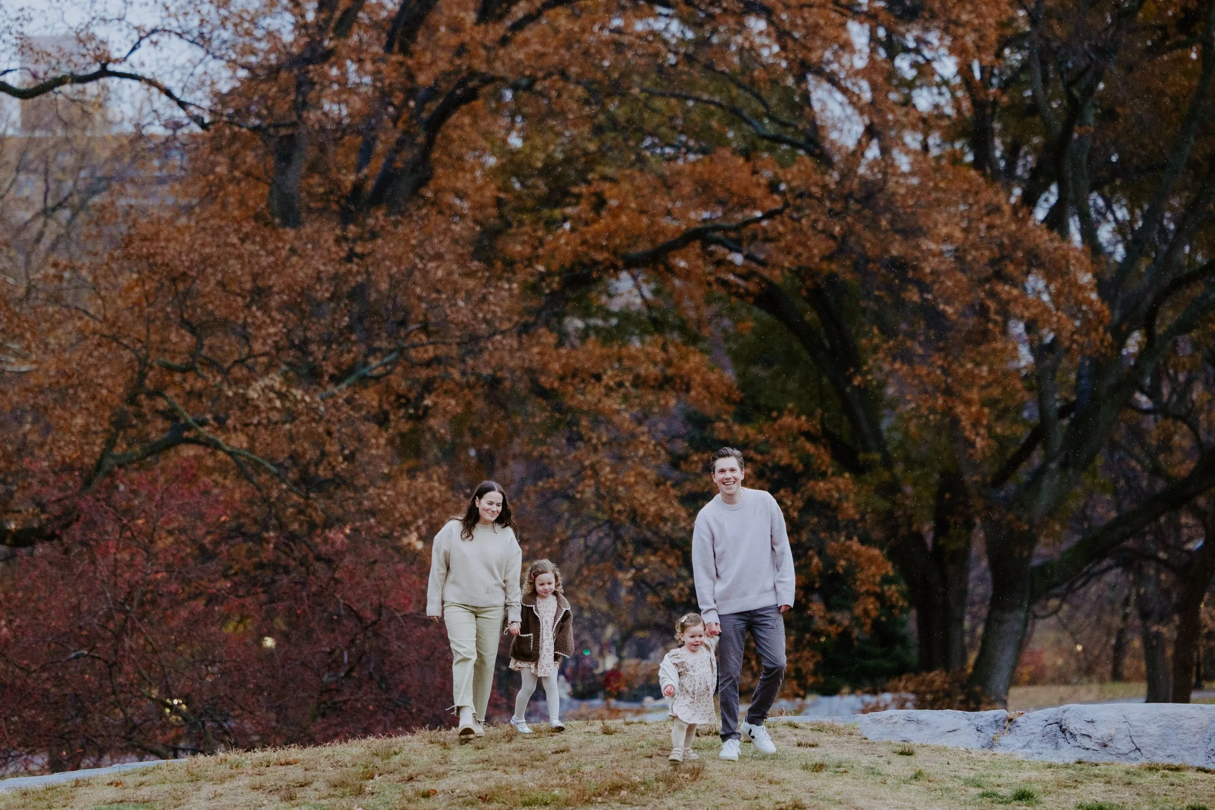 A family of four walking outdoors in a park during fall, with colorful autumn trees in the background.