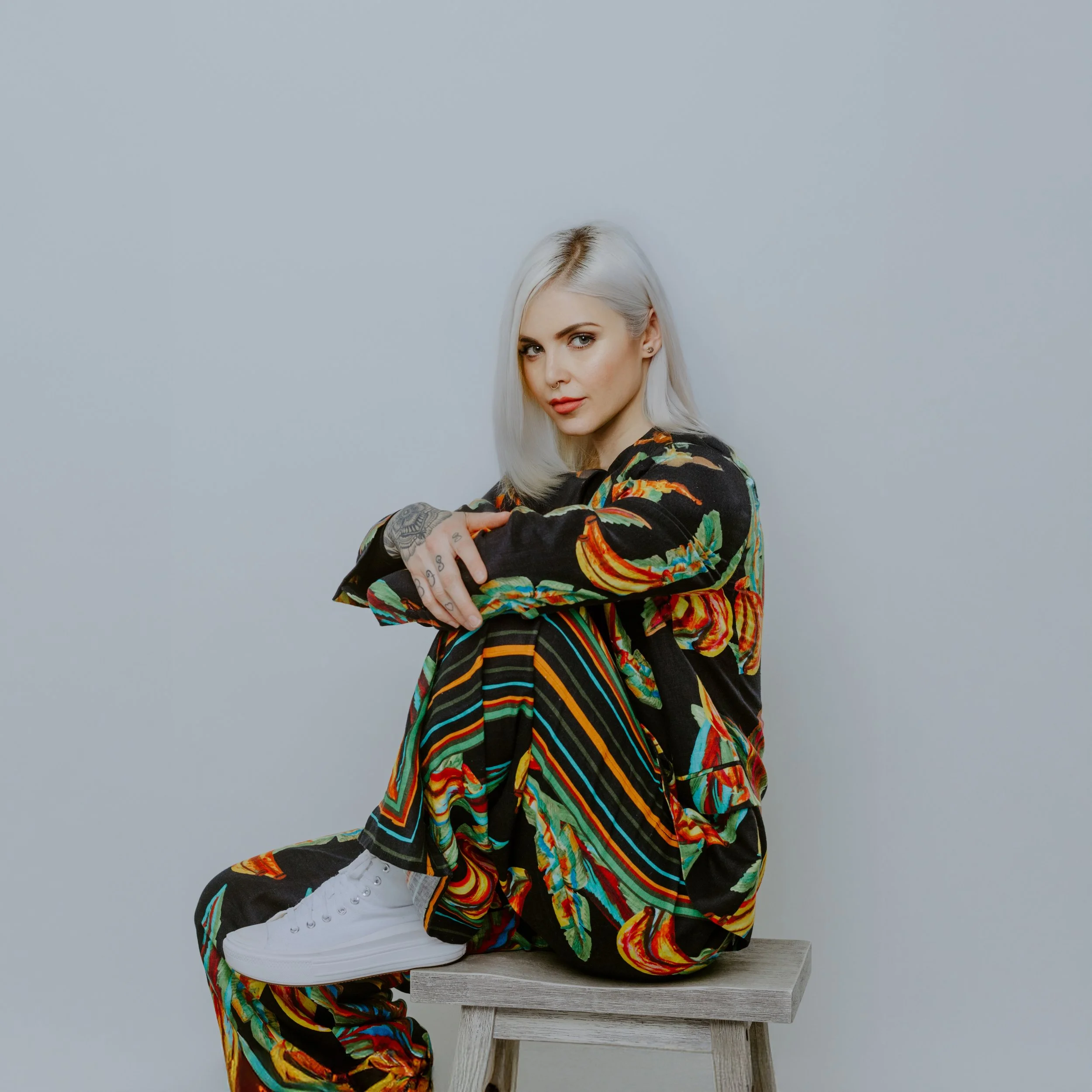 A woman with platinum blonde hair sitting on a wooden stool against a plain light gray background, wearing a colorful floral and striped patterned outfit and white sneakers.
