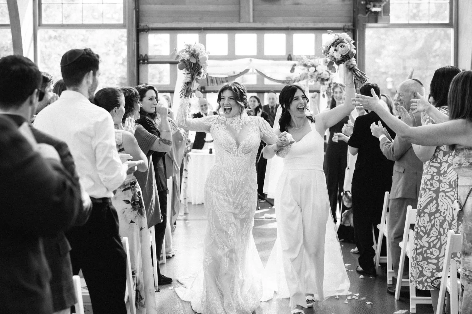 Black and white photo of a wedding celebration showing two women, one in a lace wedding dress and the other in white, happily walking through a decorated aisle, surrounded by guests reaching out for her.