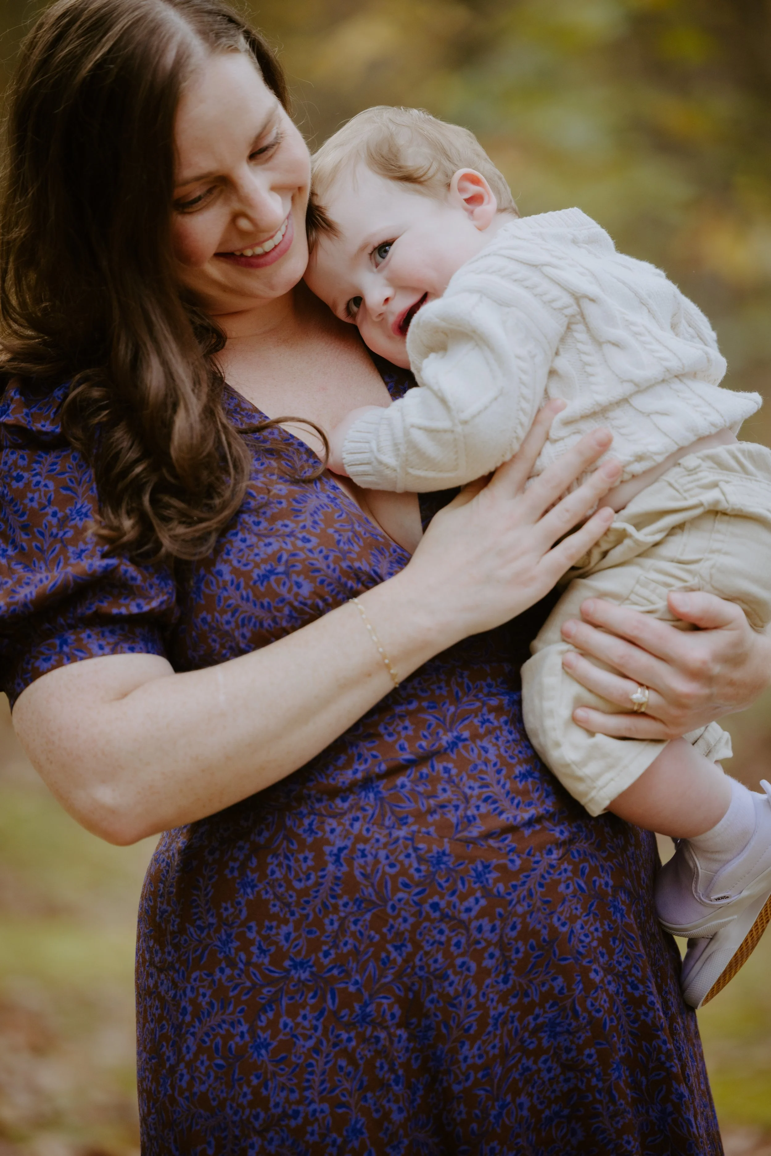 A woman with brown hair and a floral dress holding a smiling young child in an outdoor setting with autumn leaves.
