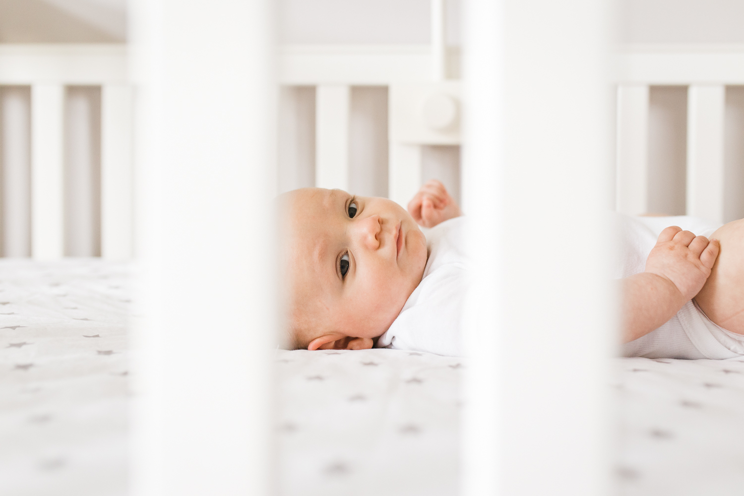 candid moment - Newborn baby boy in a white onesie looking through the side of his crib {San Francisco in-home lifestyle newborn photographer}