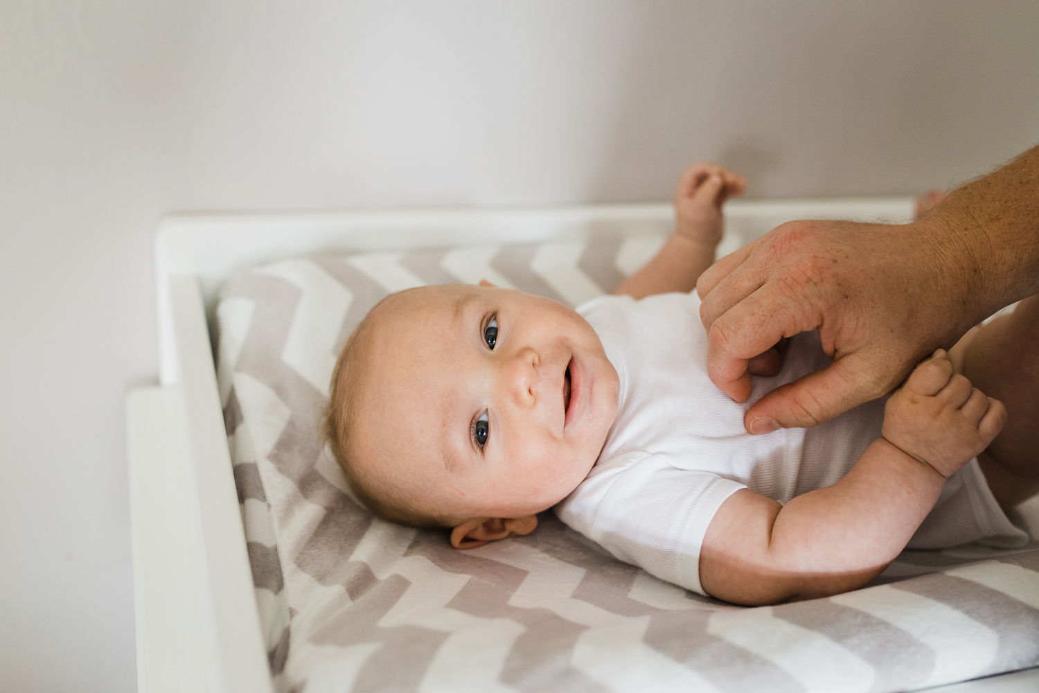 Newborn baby boy looking at the camera and smiling as he lays on his changing table with his Dad's hand on his stomach {San Francisco in-home lifestyle newborn photographer}