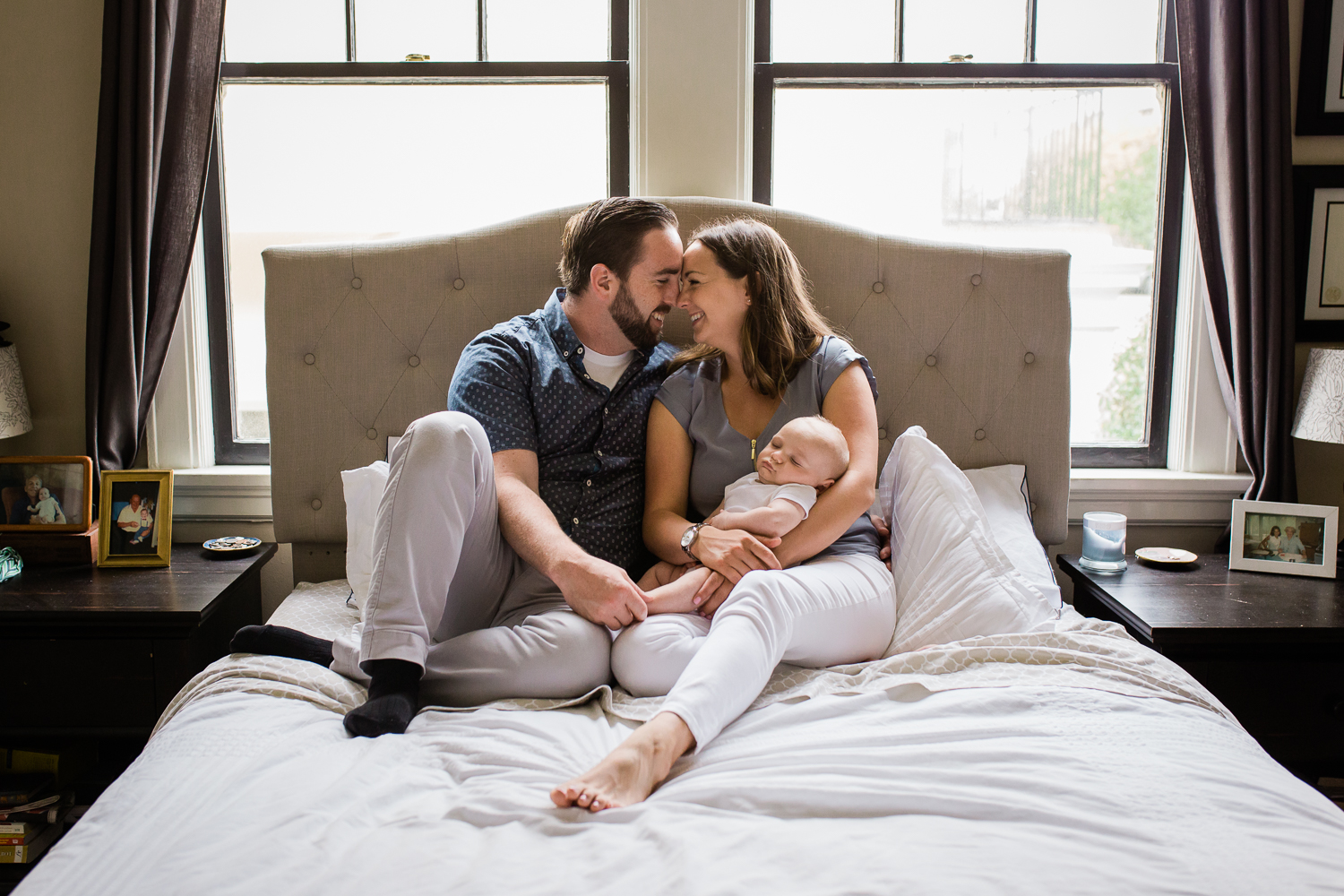 Mom holding newborn baby boy and sitting next to Dad on the bed as Mom and Dad snuggle faces together {San Francisco in-home newborn photographer}