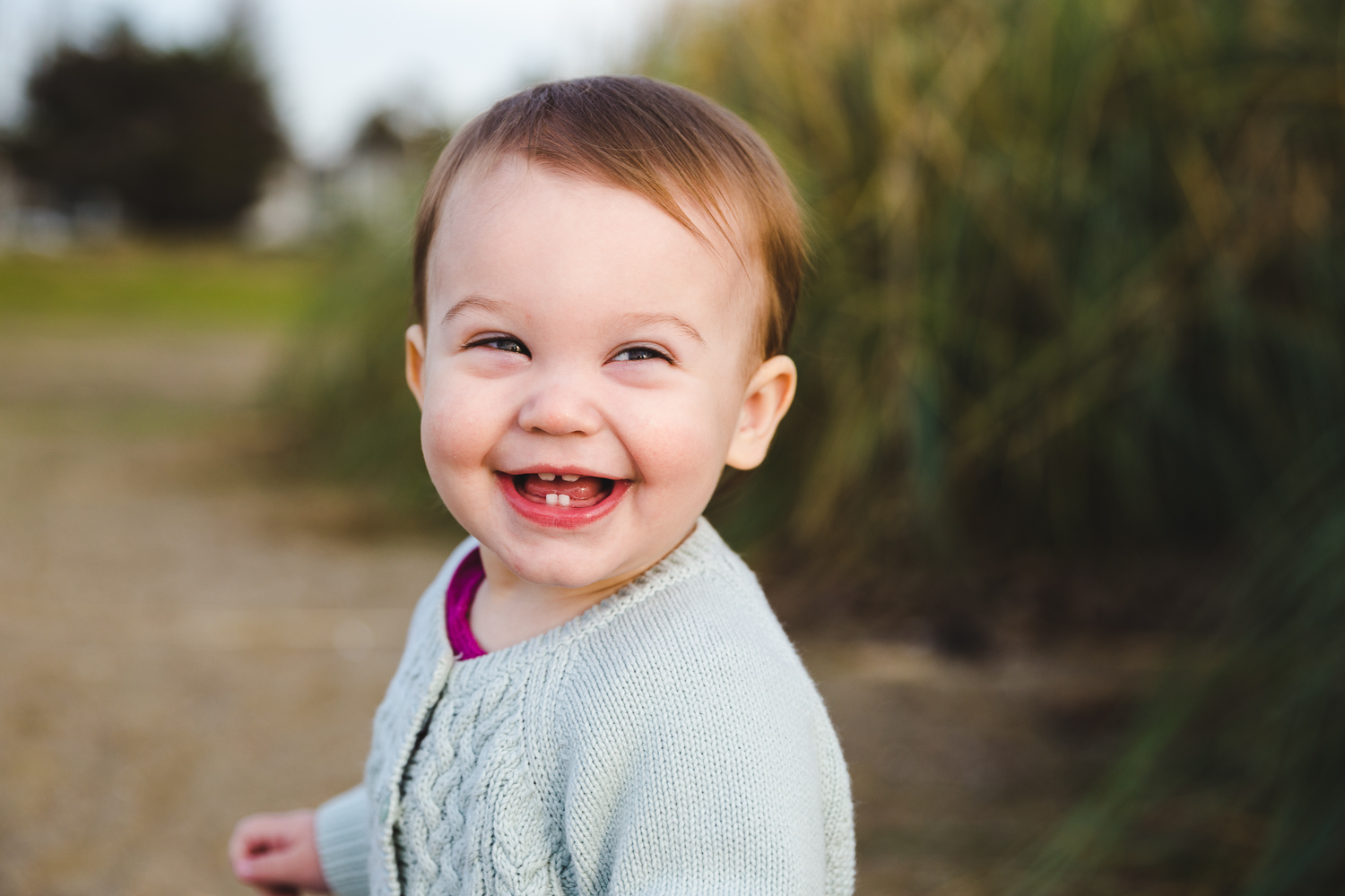 portrait of one year old baby smiling at the camera with just two bottom teeth while on the beach {East Bay Baby Photographer}