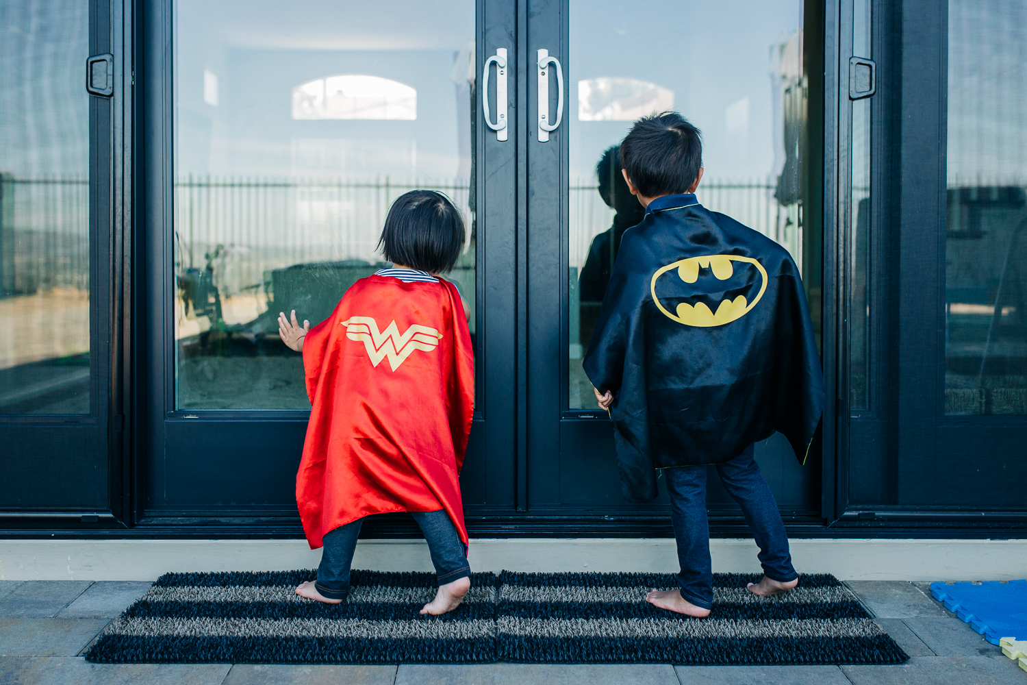 the backs of a boy and a girl wearing superhero capes as they look into sliding glass doors | East Bay Lifestyle Family Photographer