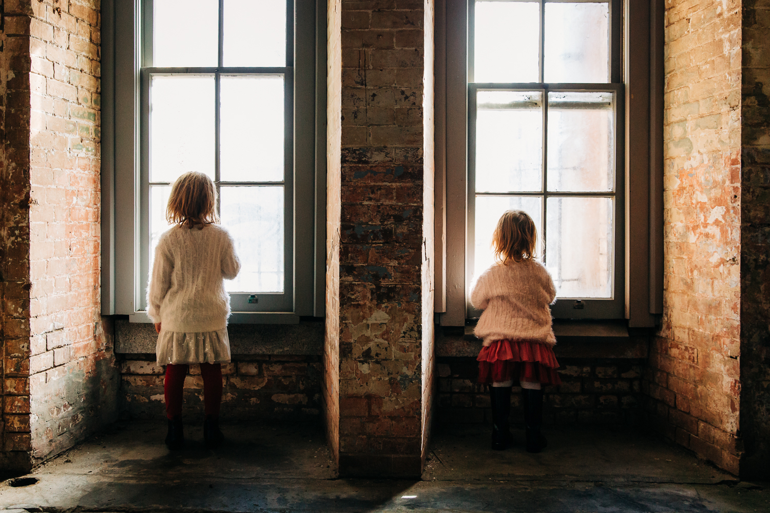 the back of two young sisters standing side next to each other looking out two separate windows  {San Francisco Family Photographer}