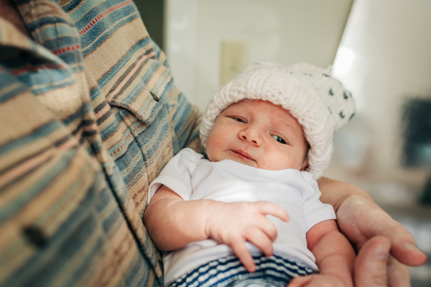 one week old baby boy in dad's arms looking towards camera while wearing a hat | SF Newborn Photographer