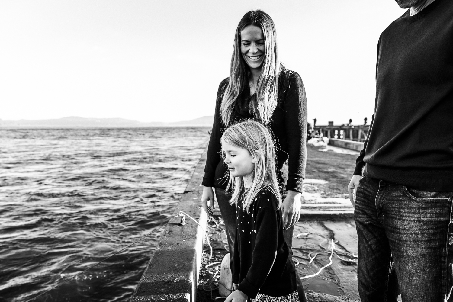 black and white shot of mom looking down at her daughter and smiling as her daughter looks in the water for fish while standing on a pier | San Francisco Portrait Photographer 