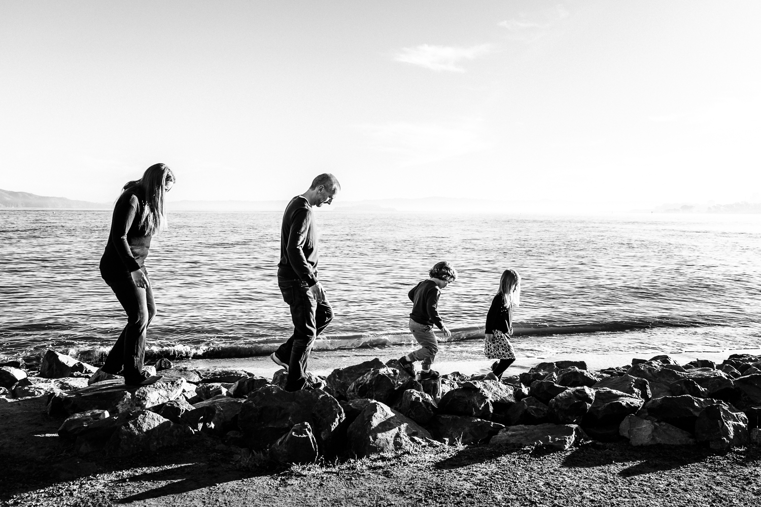 black and white photo of a family of 4 walking along rocks by the San Francisco Bay  | SF Family Photography