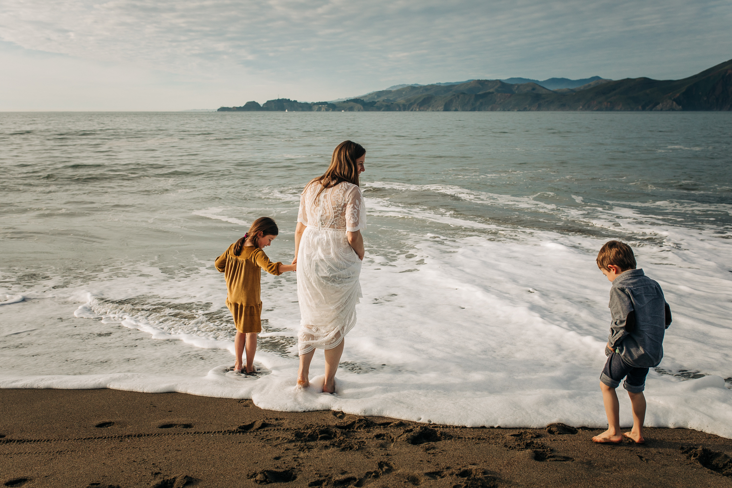 Pregnant mom playing at the beach in the water with her two children while wearing a white flowing dress | SF Maternity Photographer