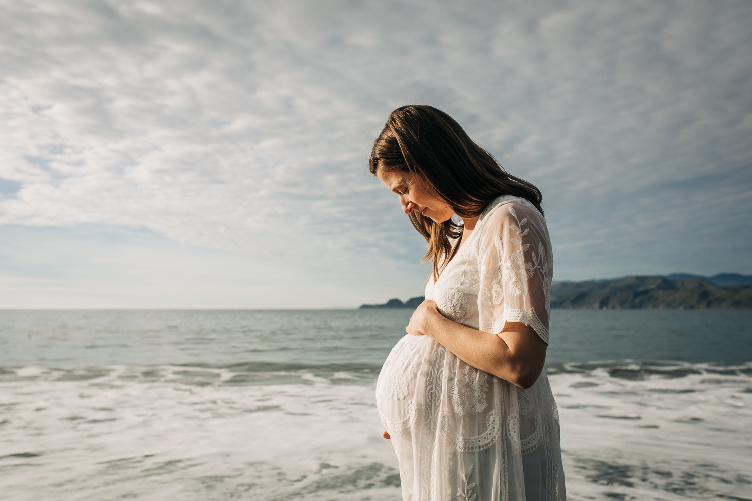 Pregnant mom looking down at her baby bump with the ocean in the background | Oakland Maternity Photographer