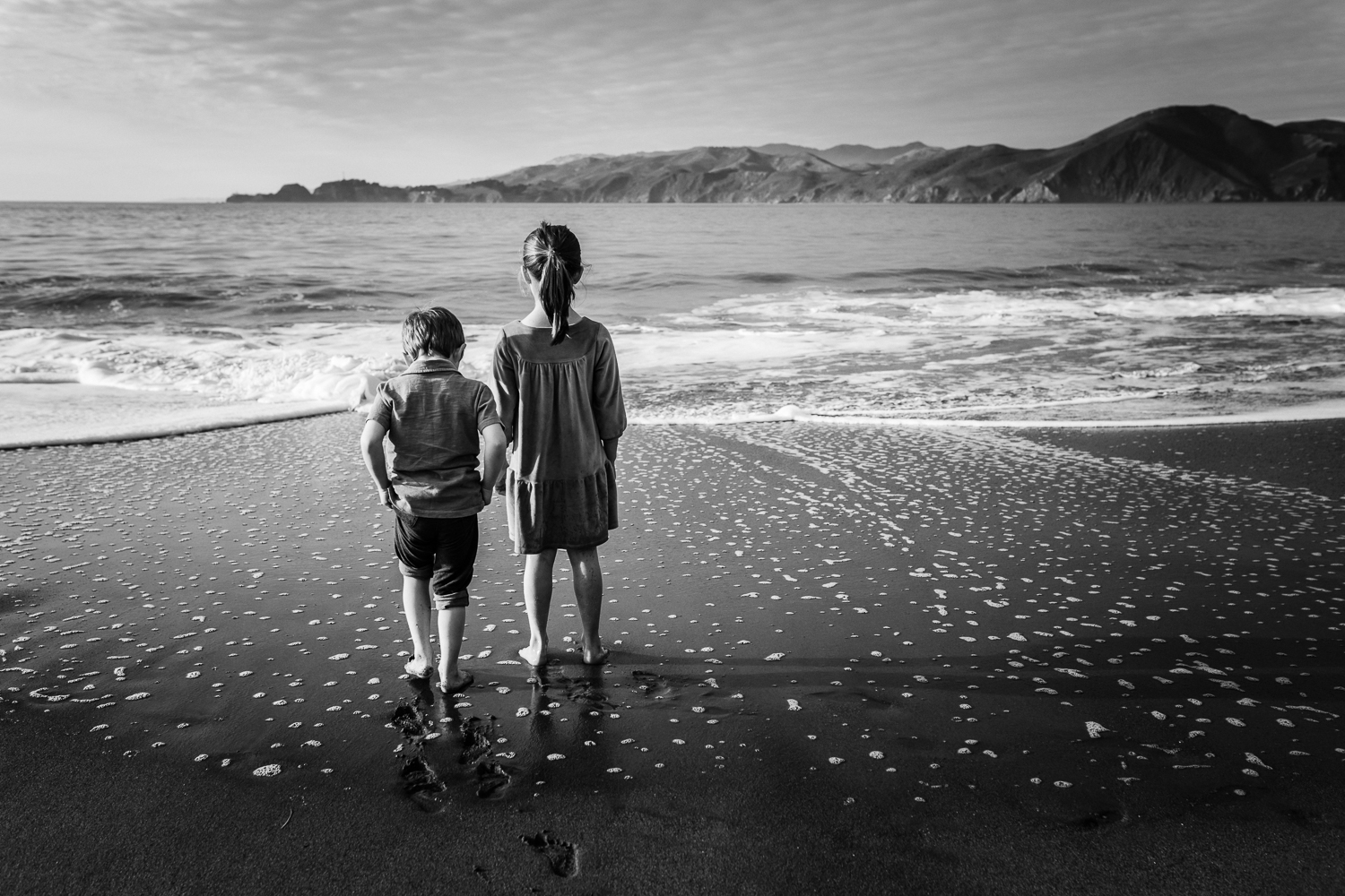 black and white photo taken from behind of two small children looking out at the ocean while standing on the beach | Bay Area Family Photographer