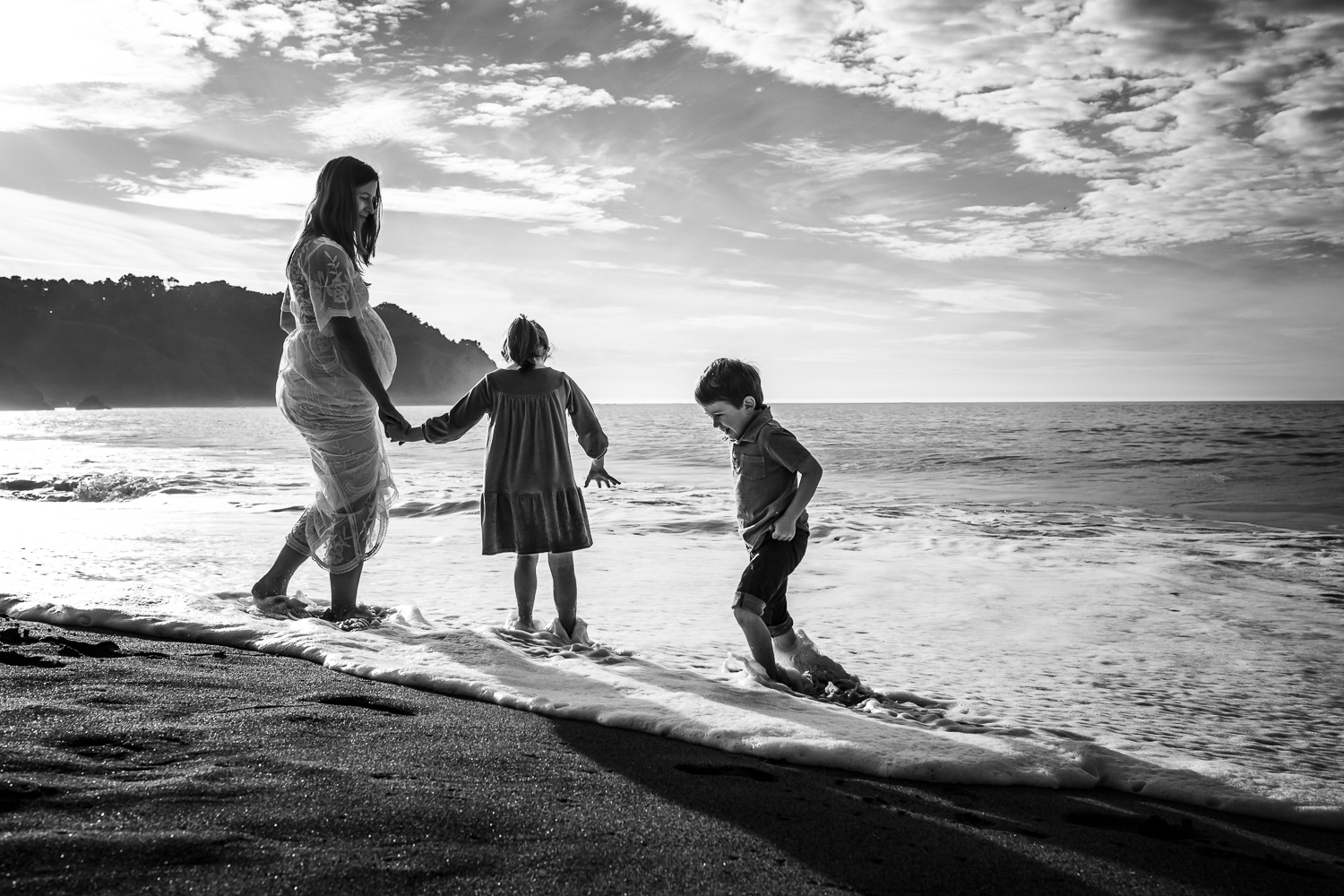 black and white photo of two small children playing in the water at the beach with their pregnant mother who is wearing a long white dress | SF Maternity Photography