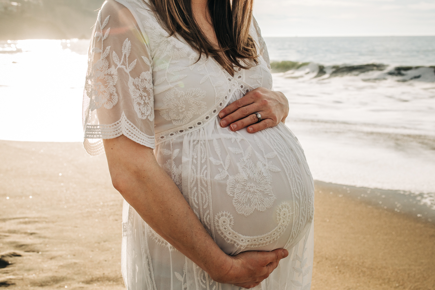 Profile photo of mother holding her baby bump while standing on the beach wearing a long white dress | Berkeley Maternity Photographer