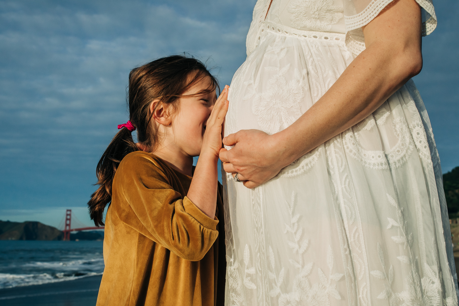 Little girl kissing her mother's baby bump at the beach with the golden gate bridge in the background | San Francisco Maternity Photographer