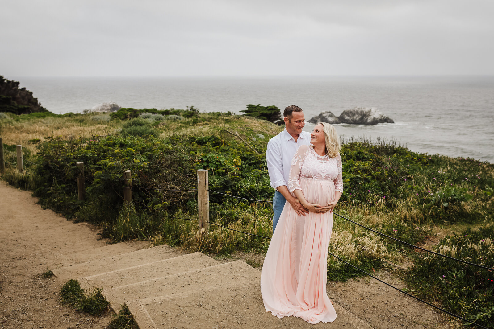 Husband standing behind pregnant wife and embracing her while standing along the coast - San Francisco Maternity Photographer 