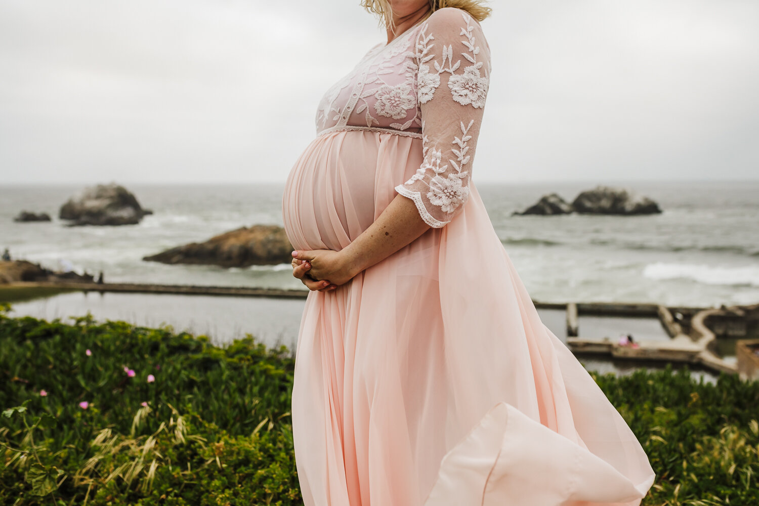 Pregnant woman holding her baby bump with dress blowing in wind standing above ocean - San Francisco Maternity Photographer